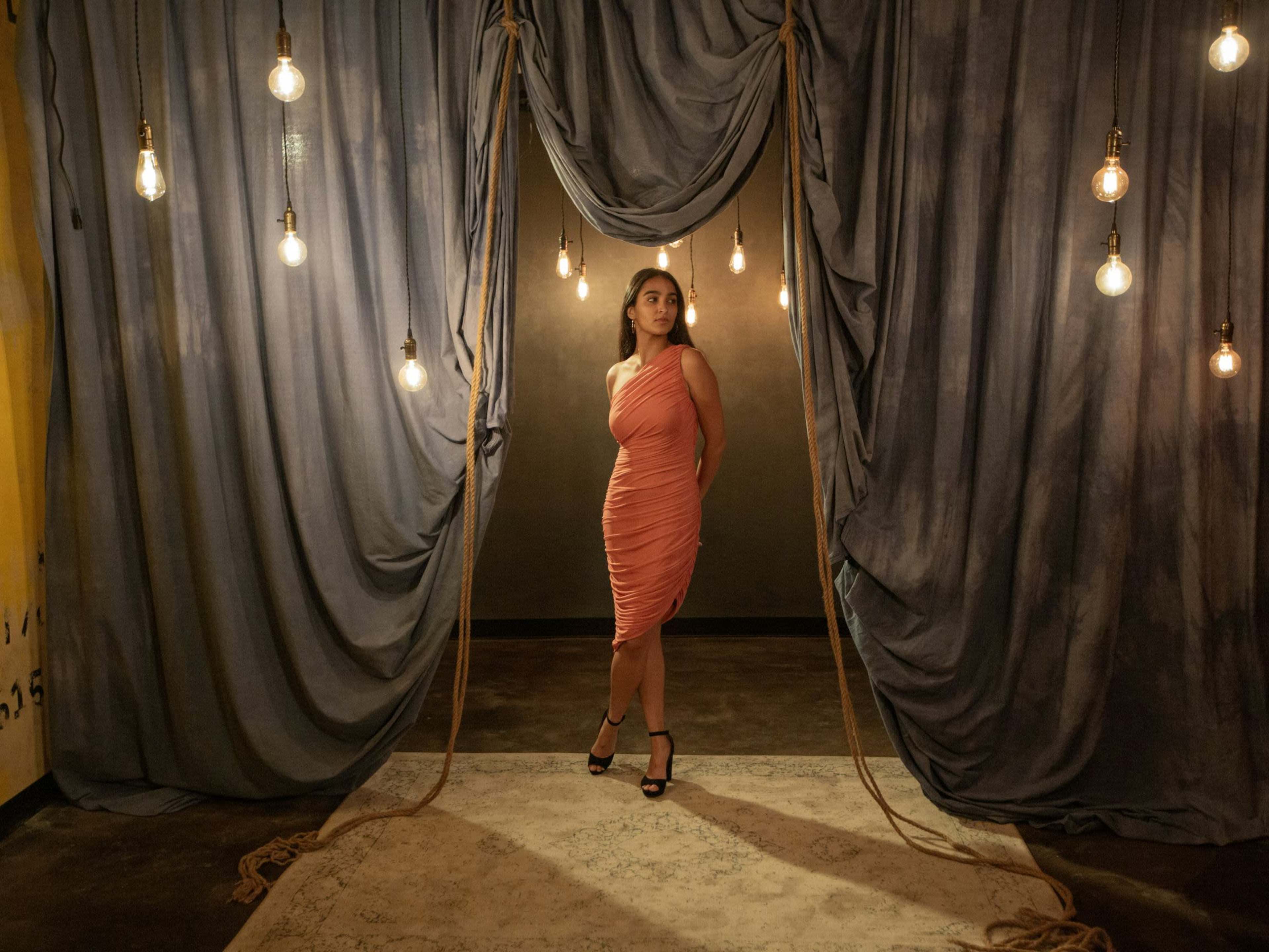 A woman in a coral dress stands center stage in front of draped fabric and suspended lights.
