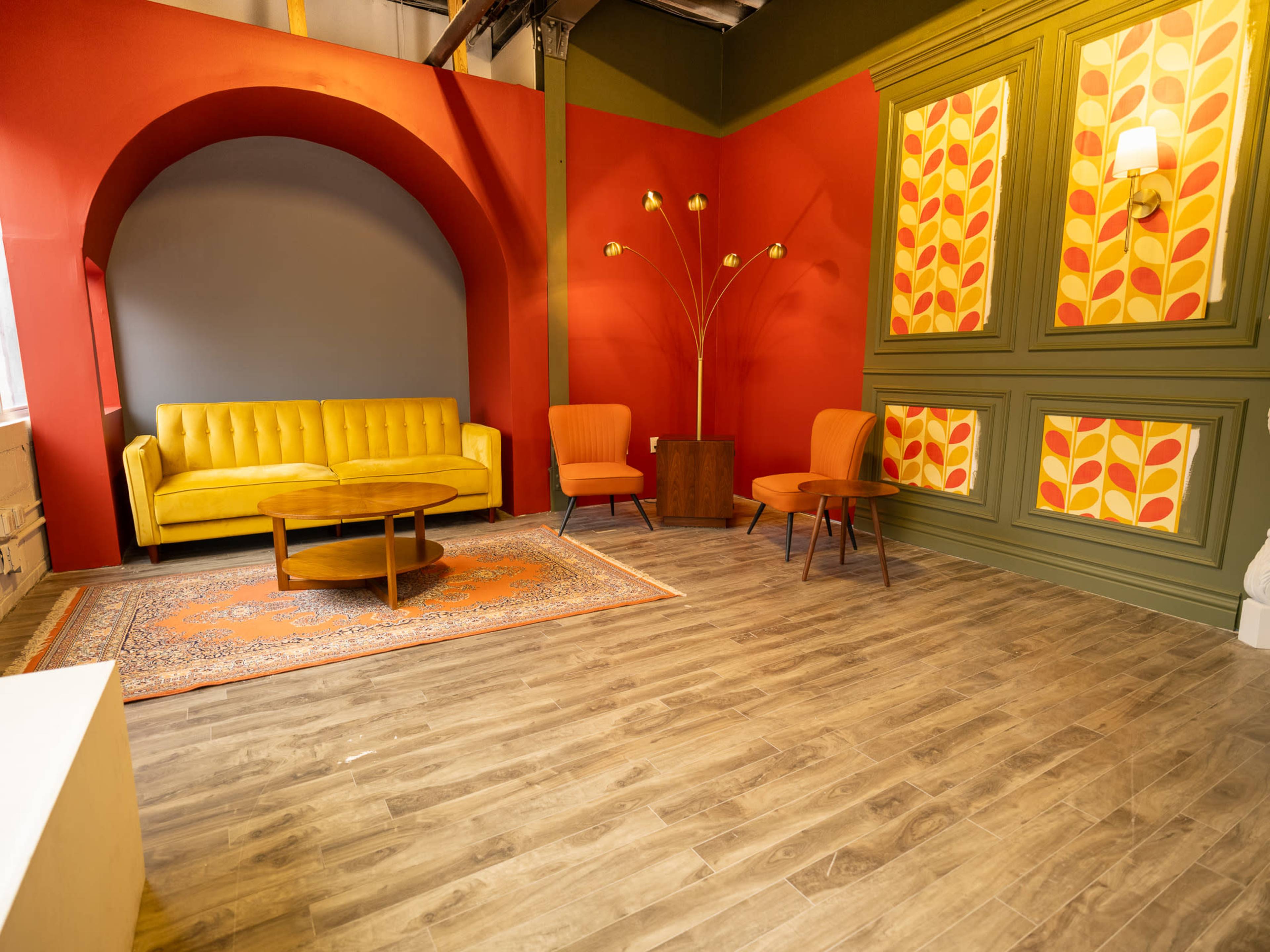 The image shows a retro-styled room with a yellow sofa, two orange chairs, a wooden coffee table, and patterned wall decor in red and green.