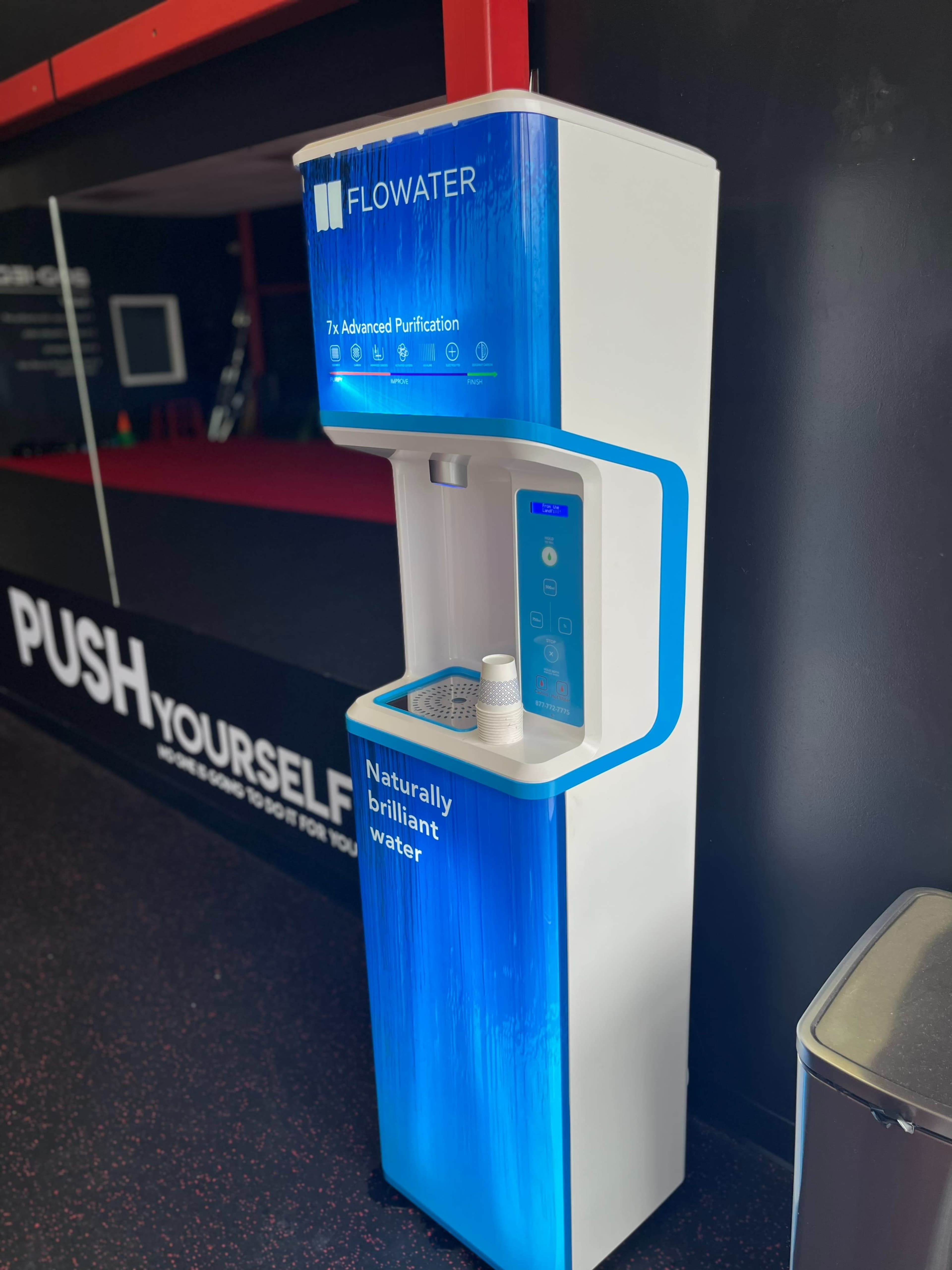 The image shows a water purification station with a sleek design, labeled "FLOWATER," featuring a button panel and a compartment for cups.