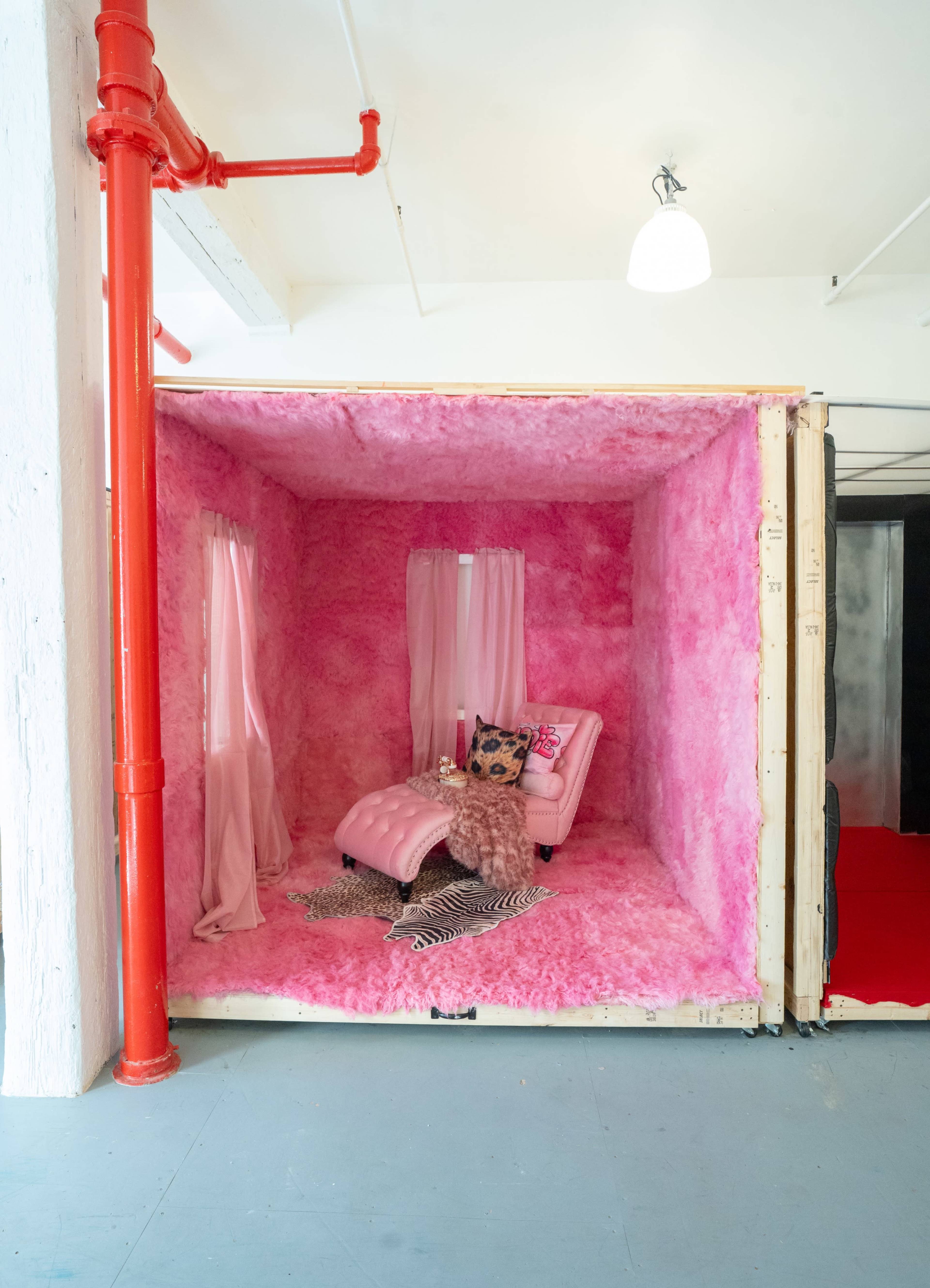 The image shows a small room filled with pink insulation material, featuring a pink chaise lounge, draped curtains, and decorative items.