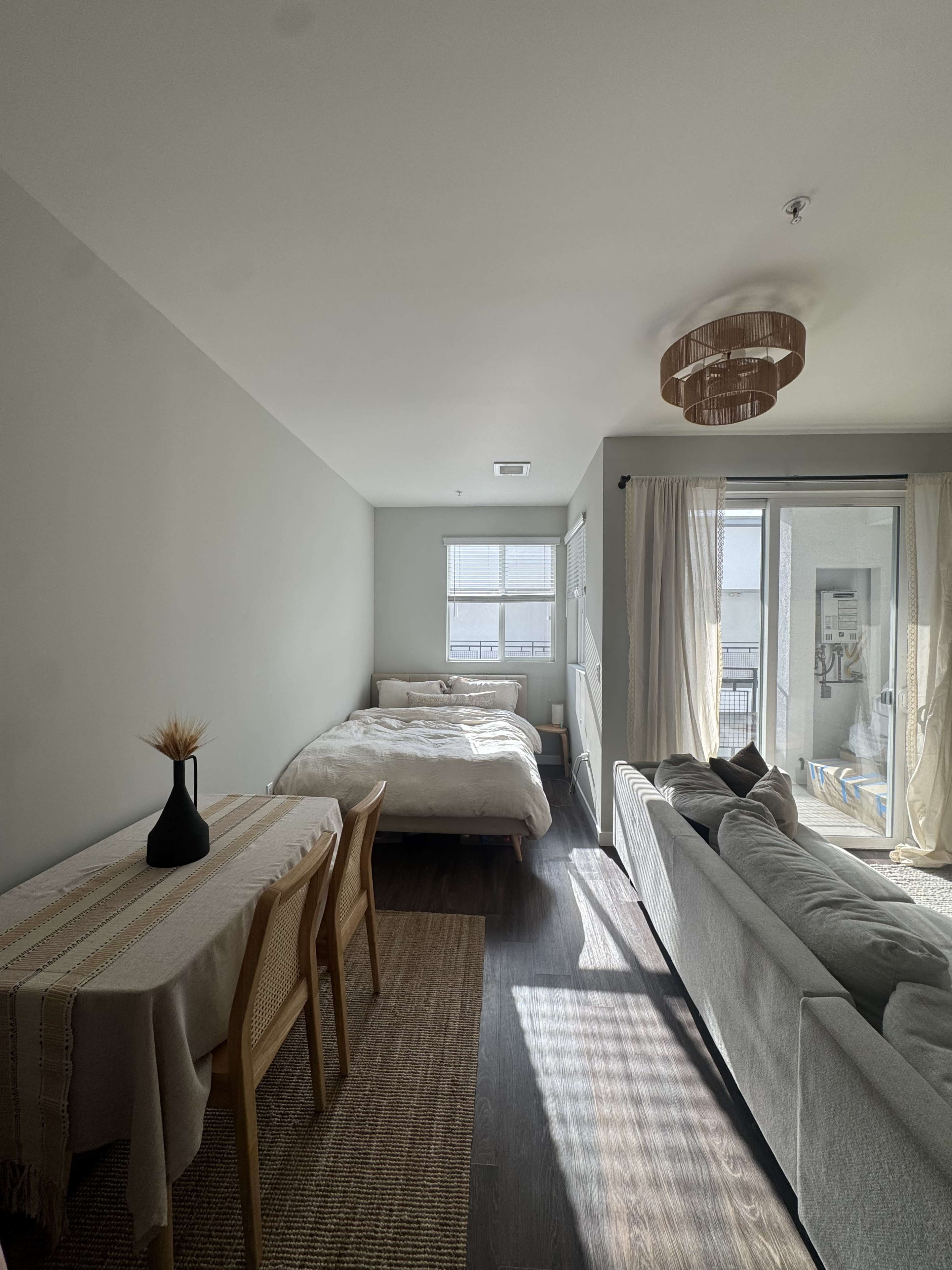 The image shows a modern studio apartment featuring a bed, dining table, and a sofa, with large windows allowing natural light to enter the space.