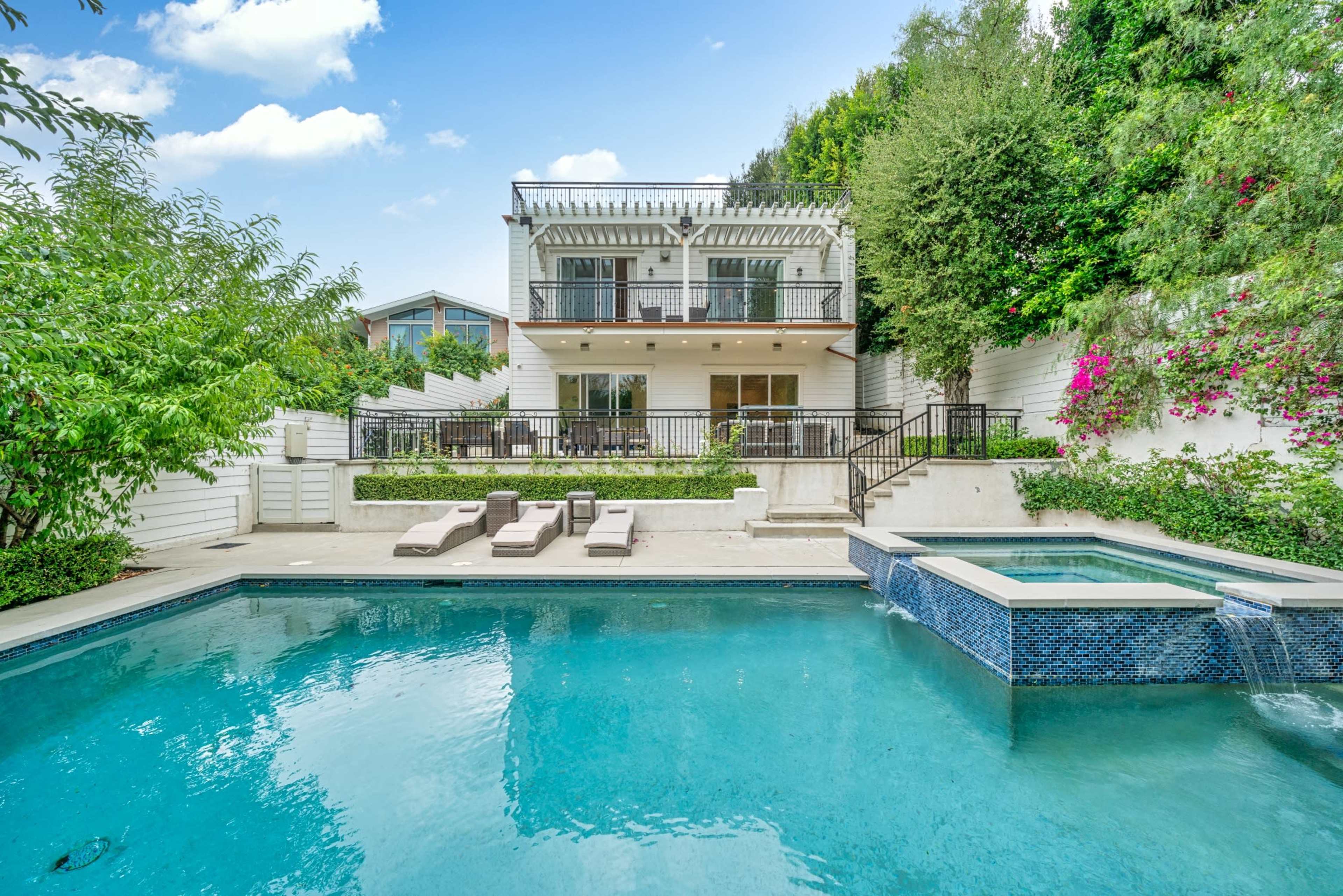 The image shows a modern two-story house with a swimming pool and lounge chairs, surrounded by greenery and flowering plants.