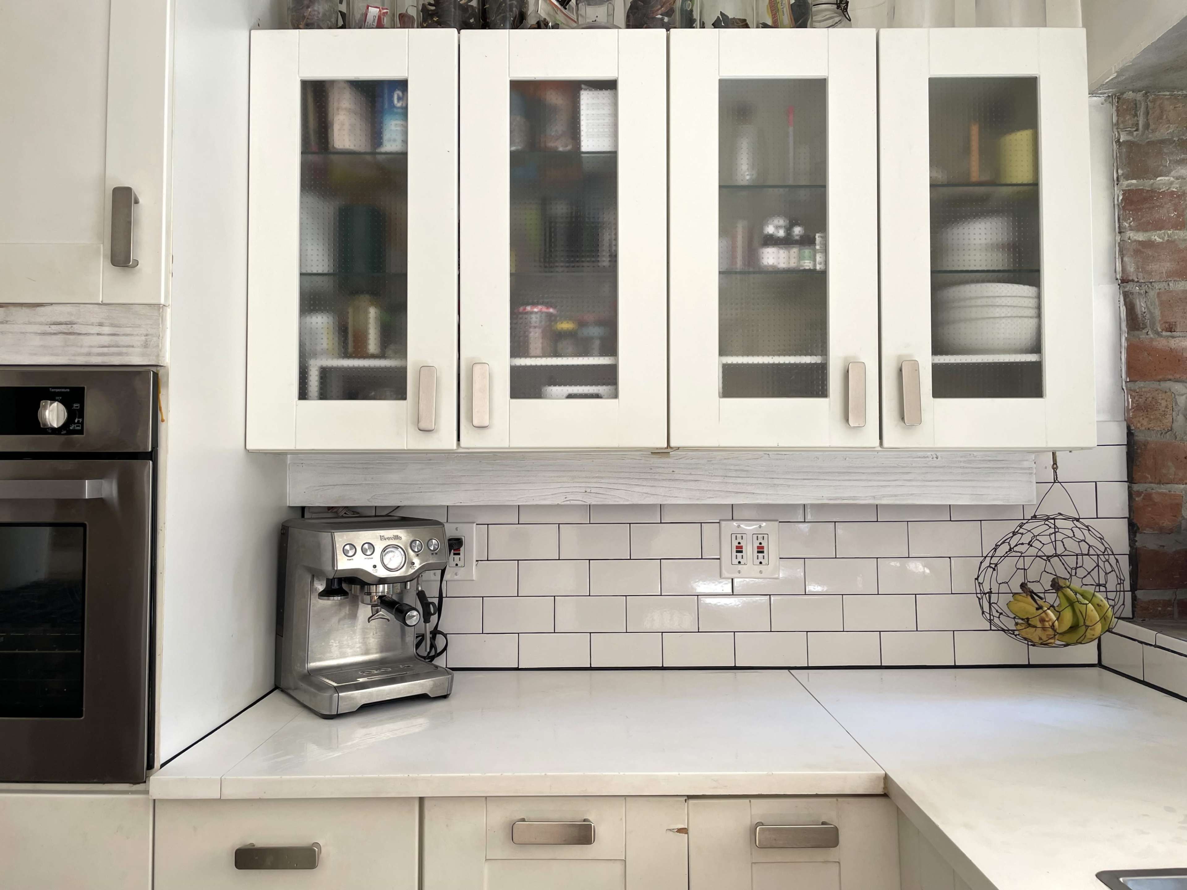 The image shows a kitchen cabinet with glass-front doors displaying various kitchen items, alongside a countertop featuring a coffee machine and a wire bowl of bananas.