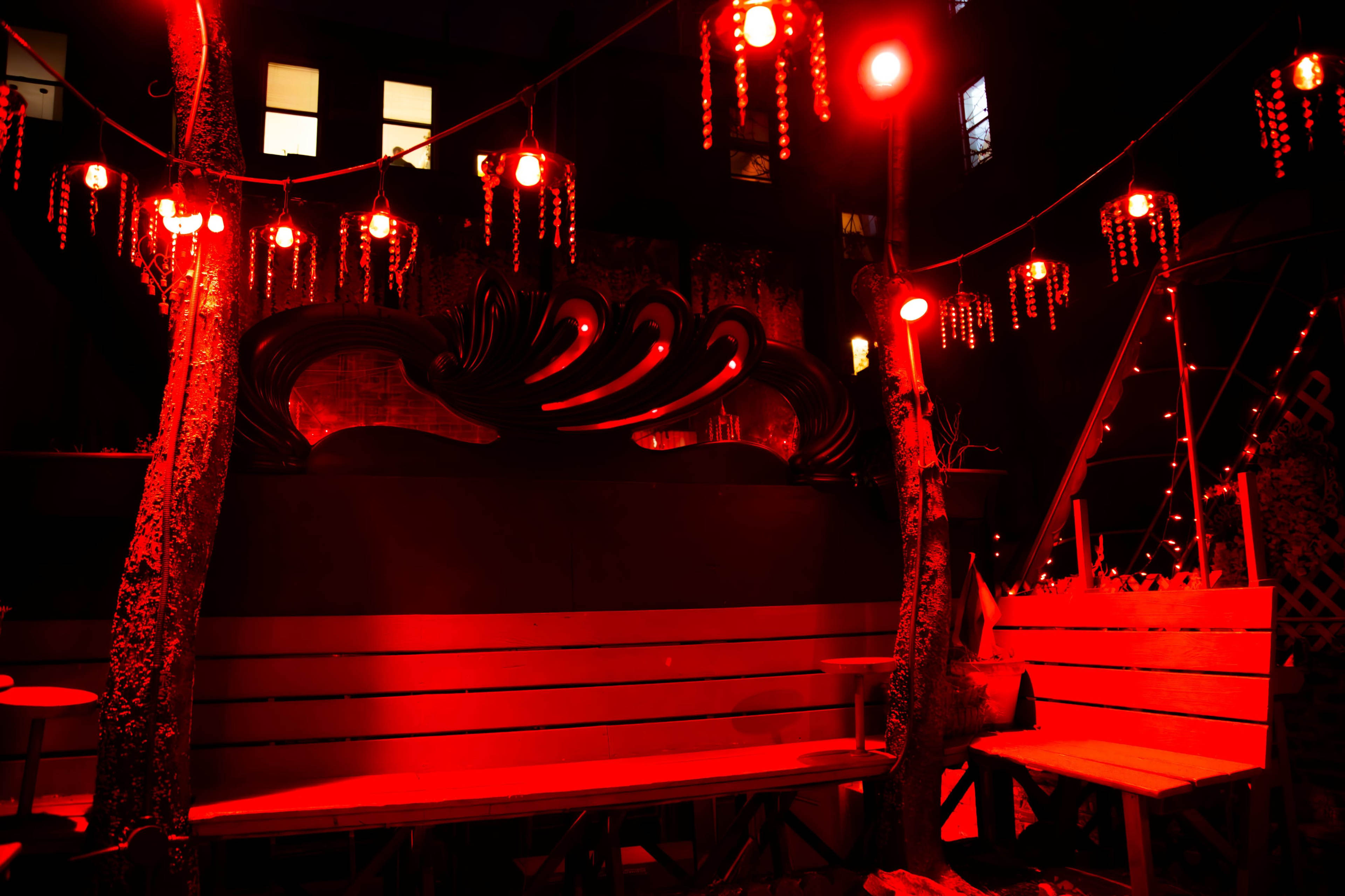 The image shows a dimly lit outdoor seating area illuminated by red lights, featuring wooden benches and decorative hanging elements.