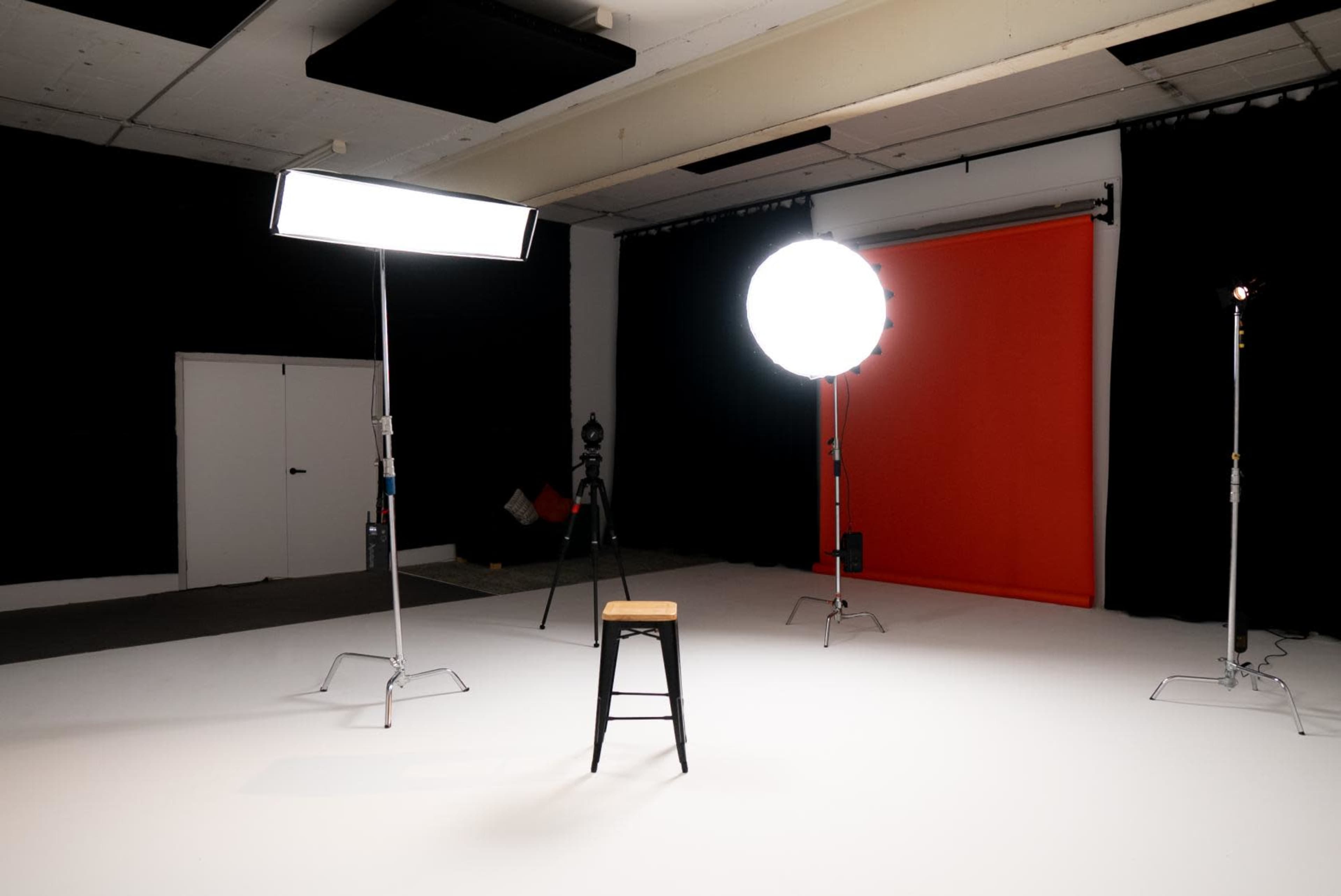A black stool is positioned in the center of an empty studio with bright lighting and an orange backdrop.