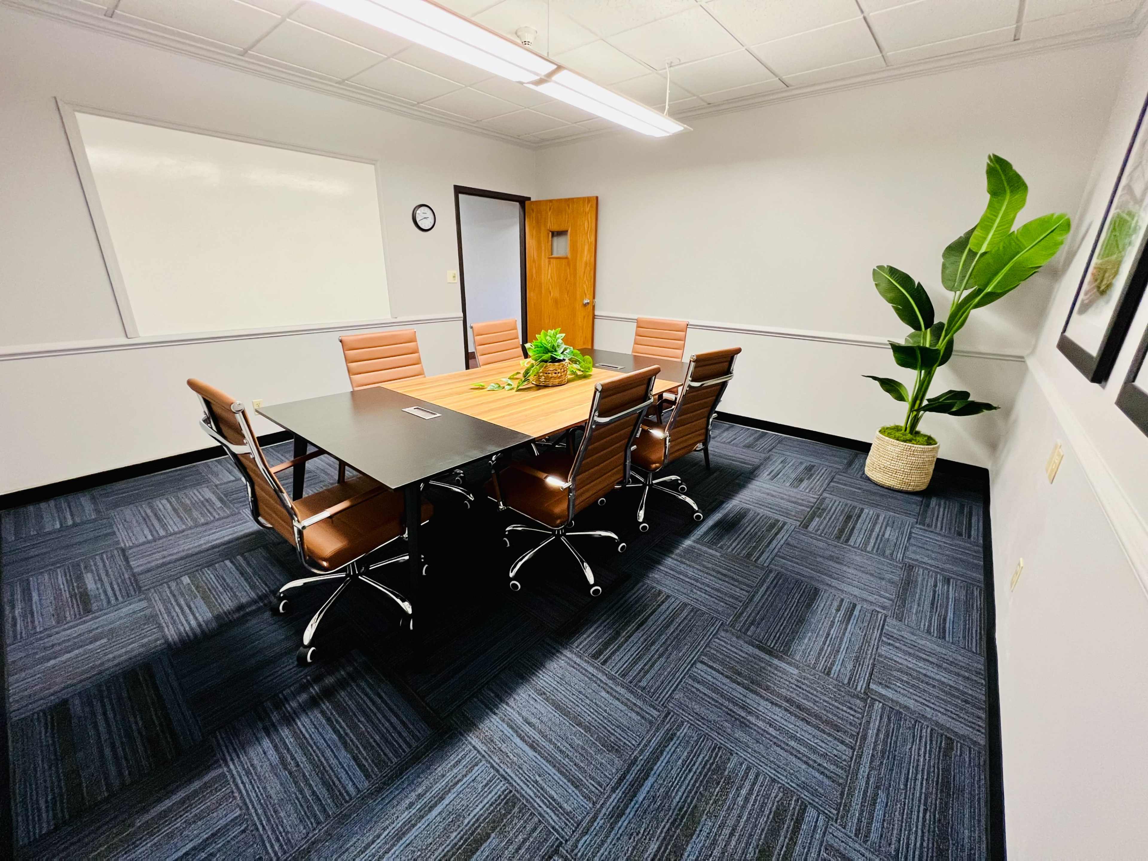 A conference room features a large table surrounded by several brown leather chairs and a potted plant in the corner.