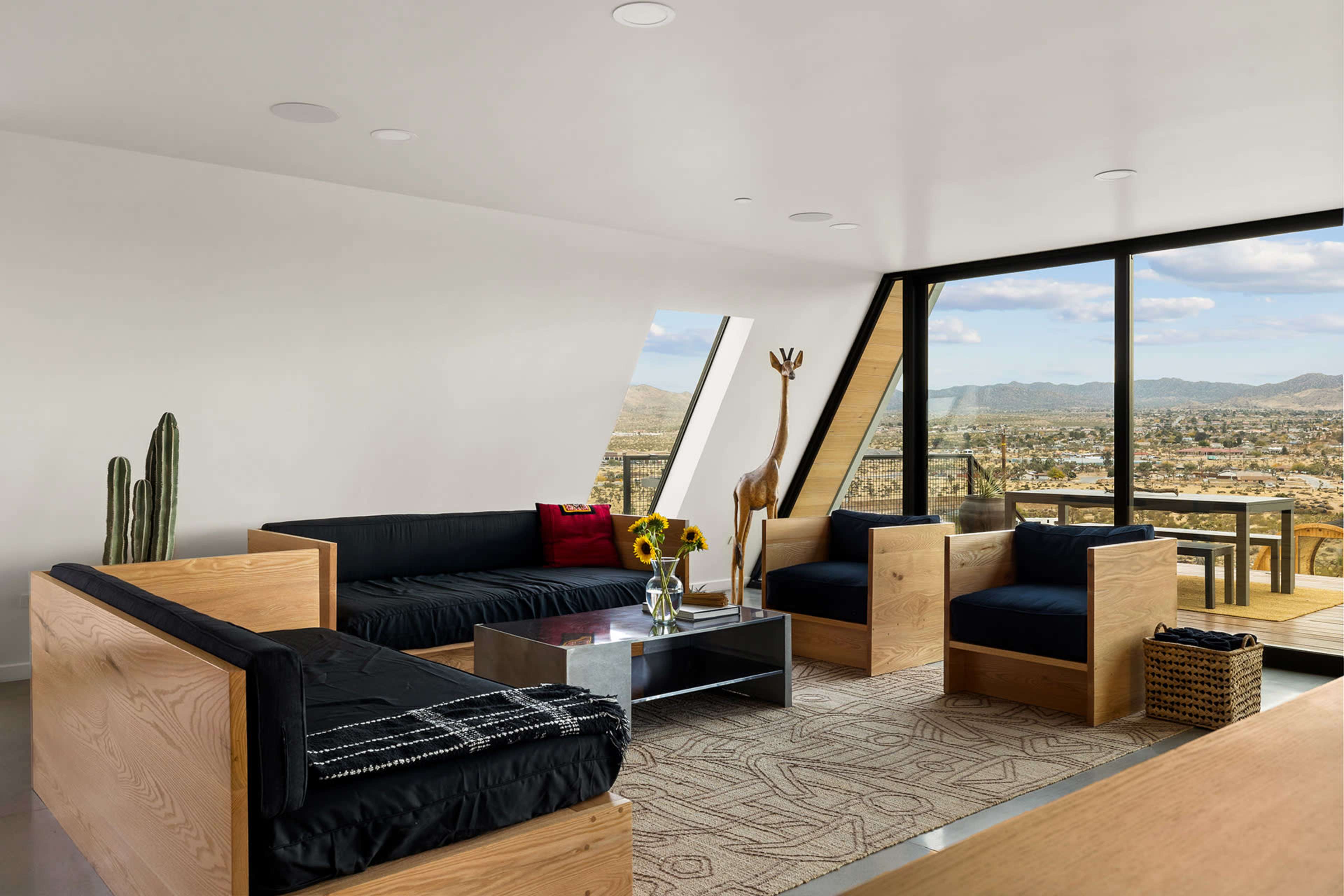A modern living room features minimalist furniture, a view of the landscape through large windows, and decorative elements like a giraffe statue and cacti.