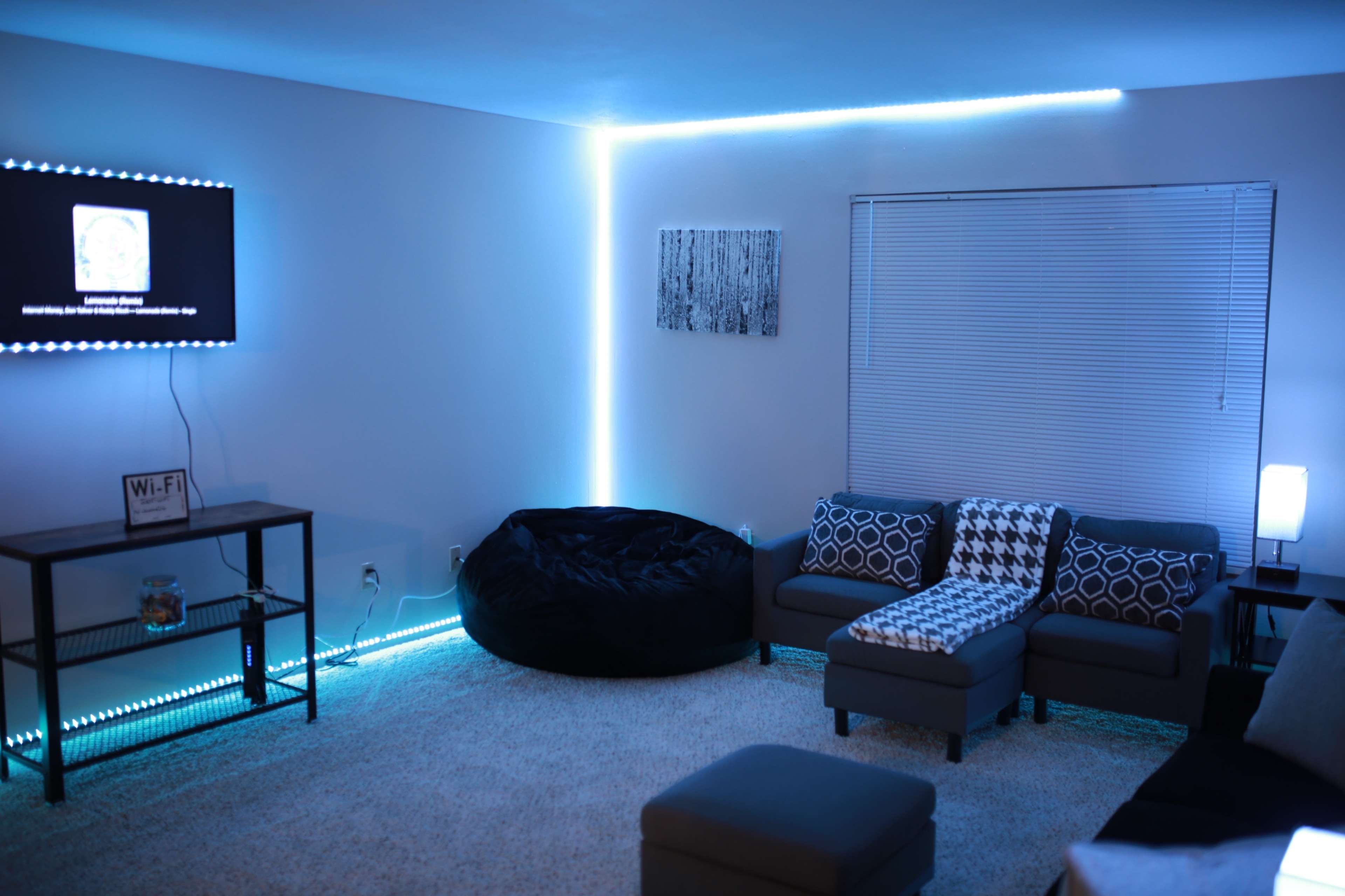 A modern living room with a sectional sofa, a black bean bag chair, and LED lights illuminating the walls, along with a TV mounted on the wall and a small table displaying a Wi-Fi sign.