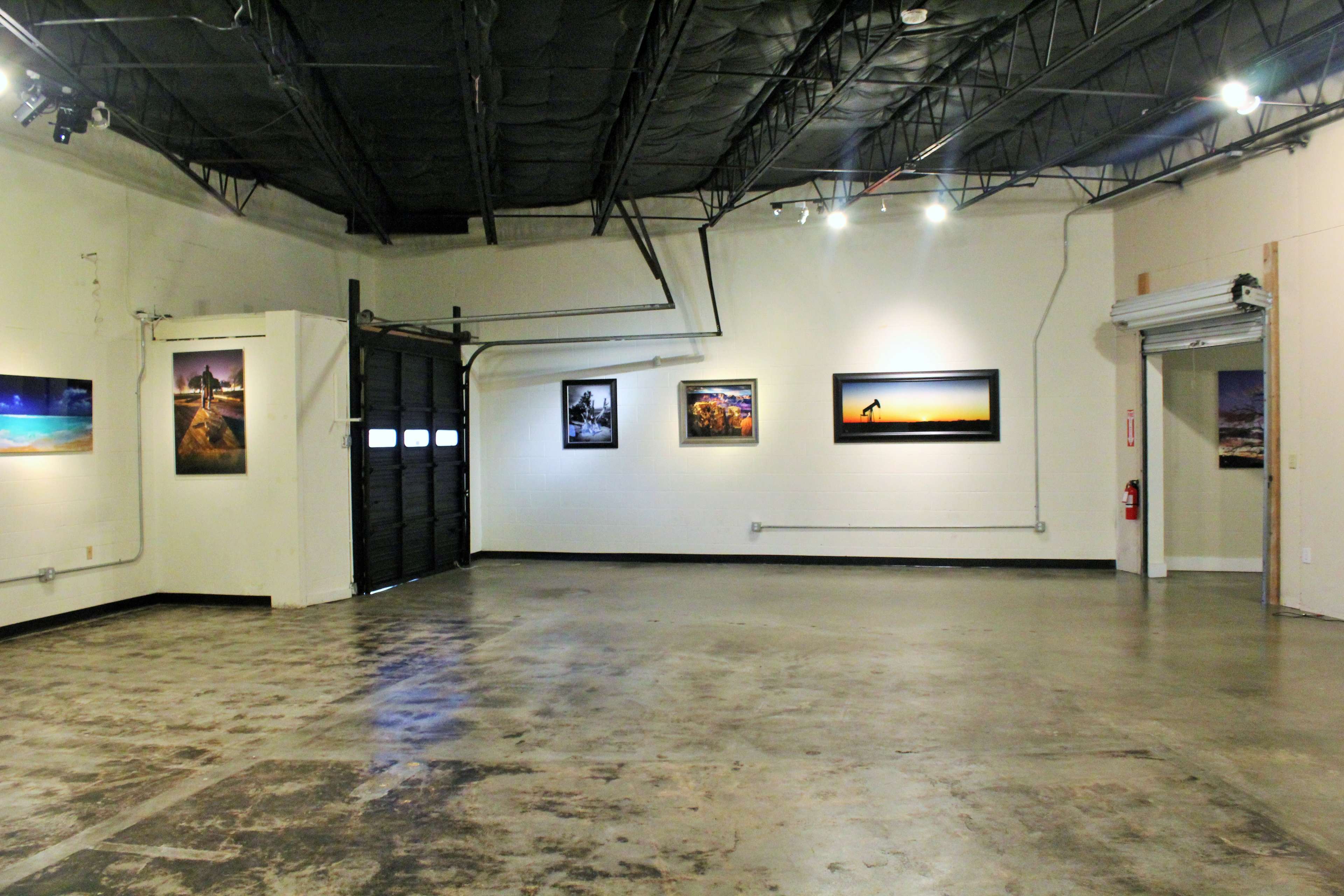 The image shows an empty art gallery with several photographs displayed on the walls and a large garage door on one side.