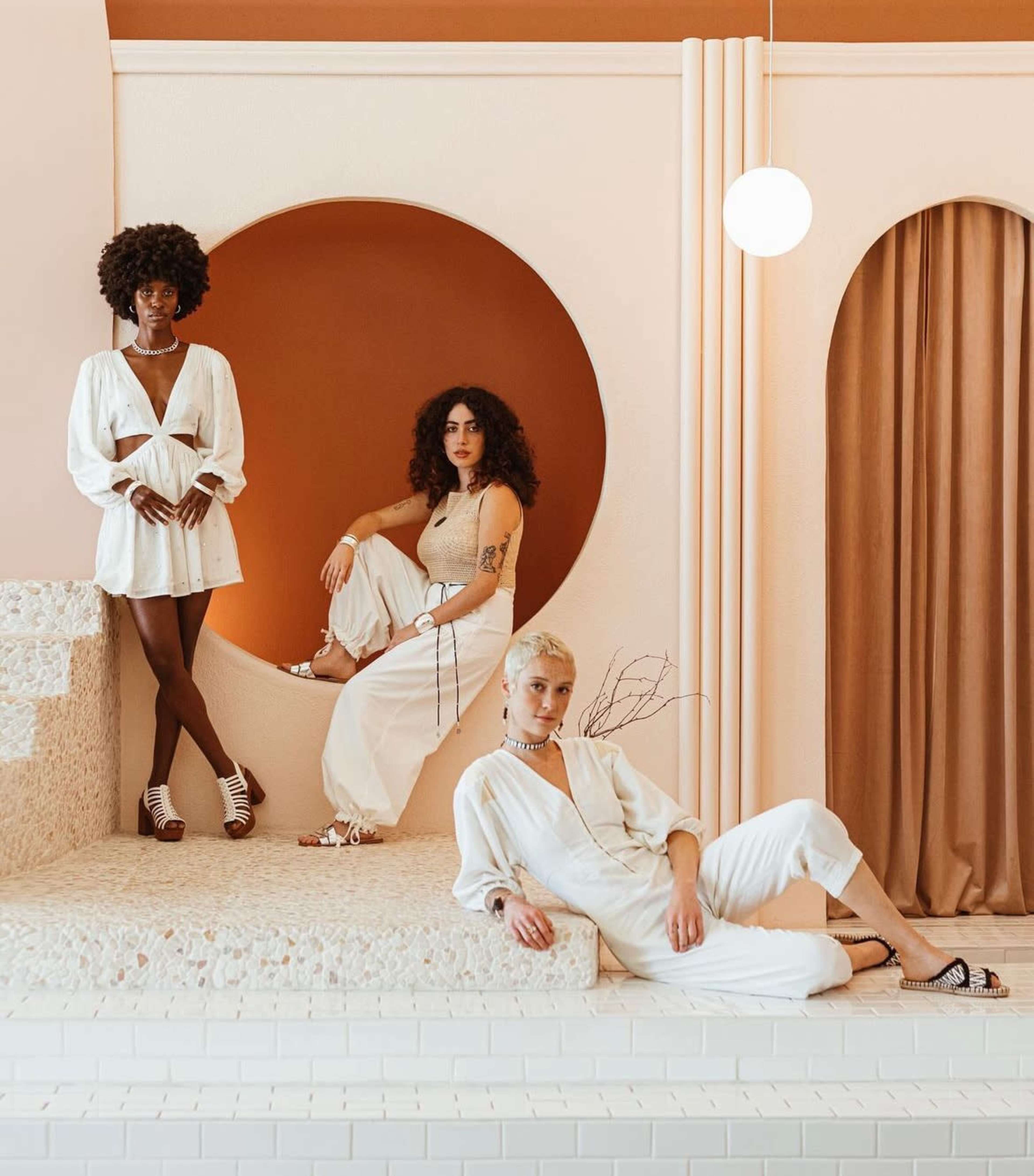 Three women in white outfits pose in a modern, geometric interior featuring a circular orange accent and cream-colored textures.