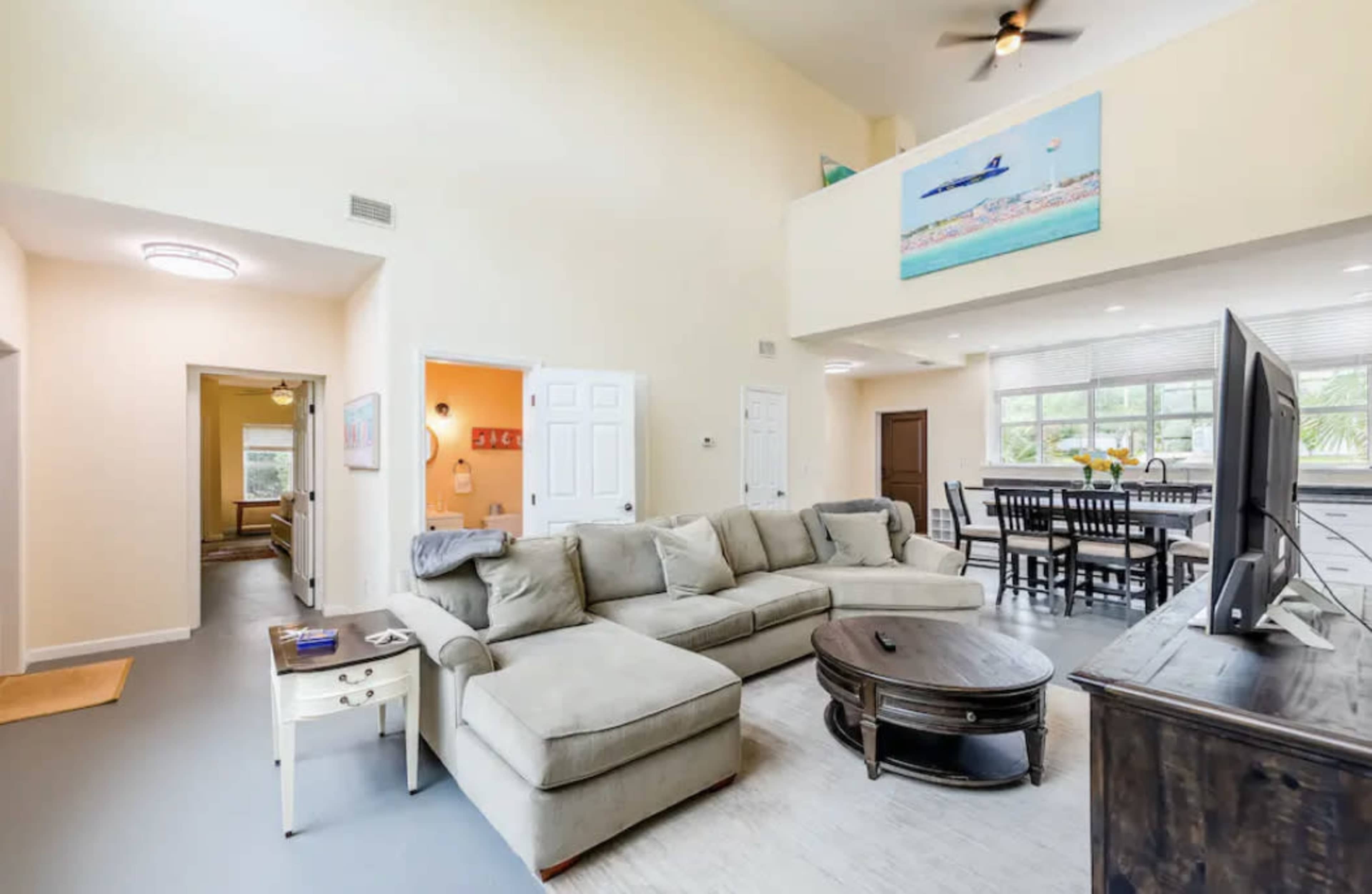 A spacious living area with a gray sectional sofa, a round coffee table, and a dining space featuring a table with chairs, all illuminated by natural light from large windows.