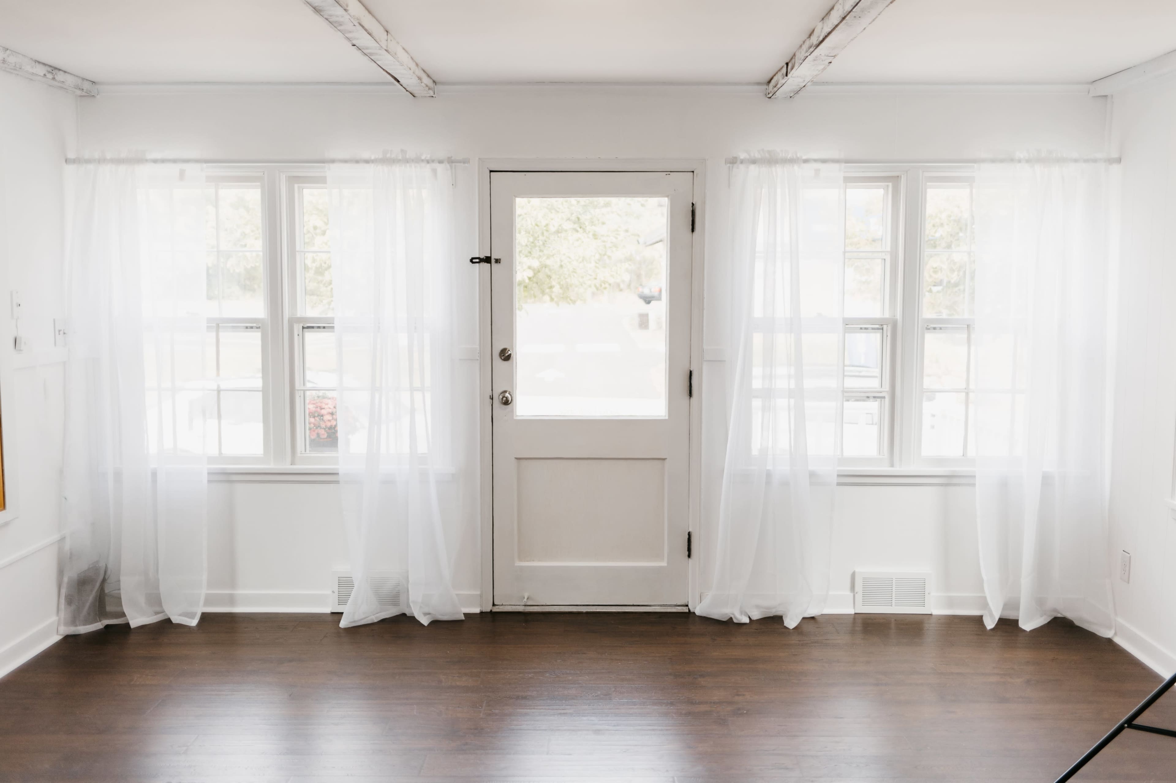 A bright, minimalist room features two windows with sheer white curtains and a white door leading outside.