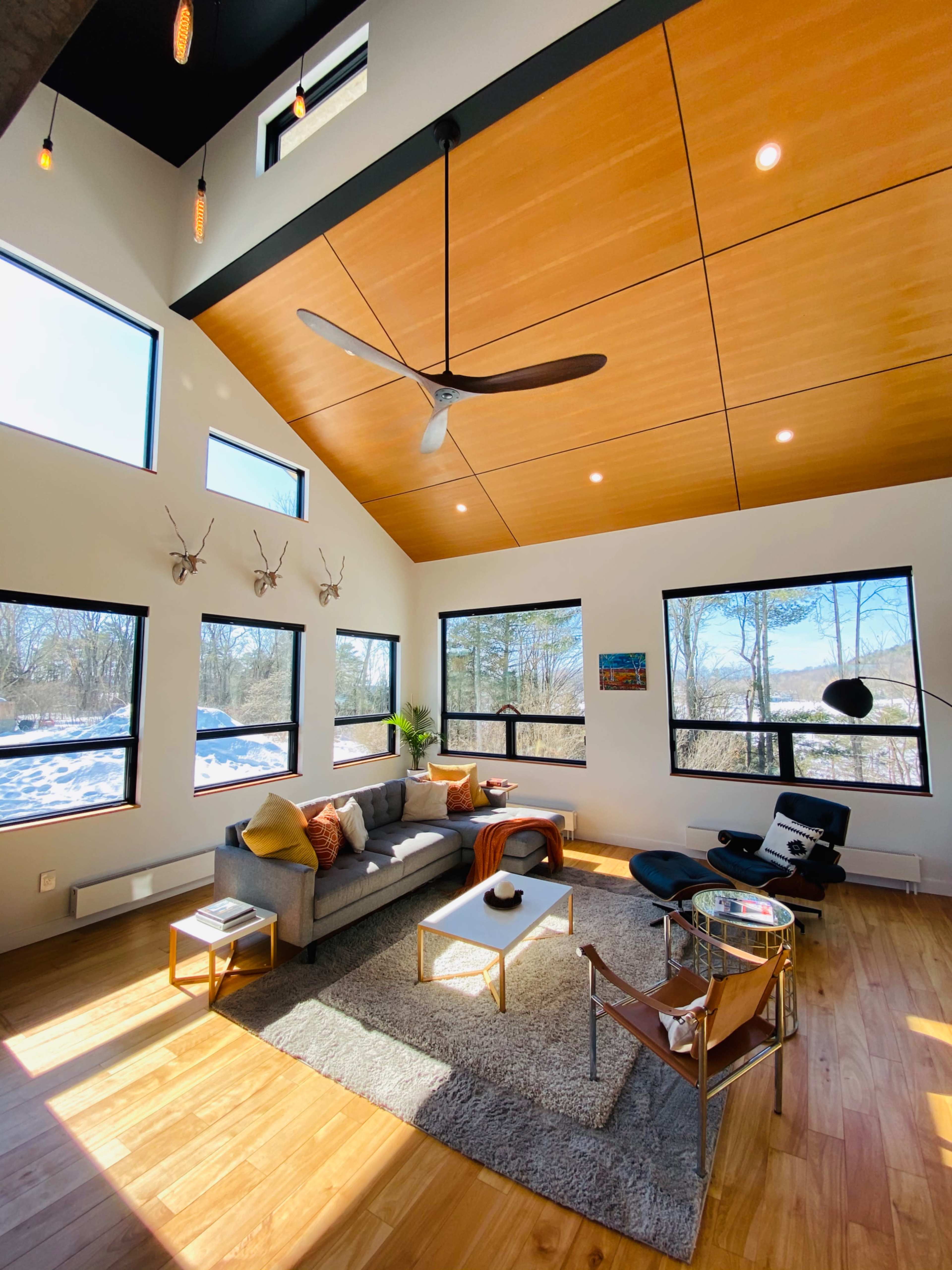 A modern living room with large windows, a ceiling fan, and stylish furniture, illuminated by natural light.