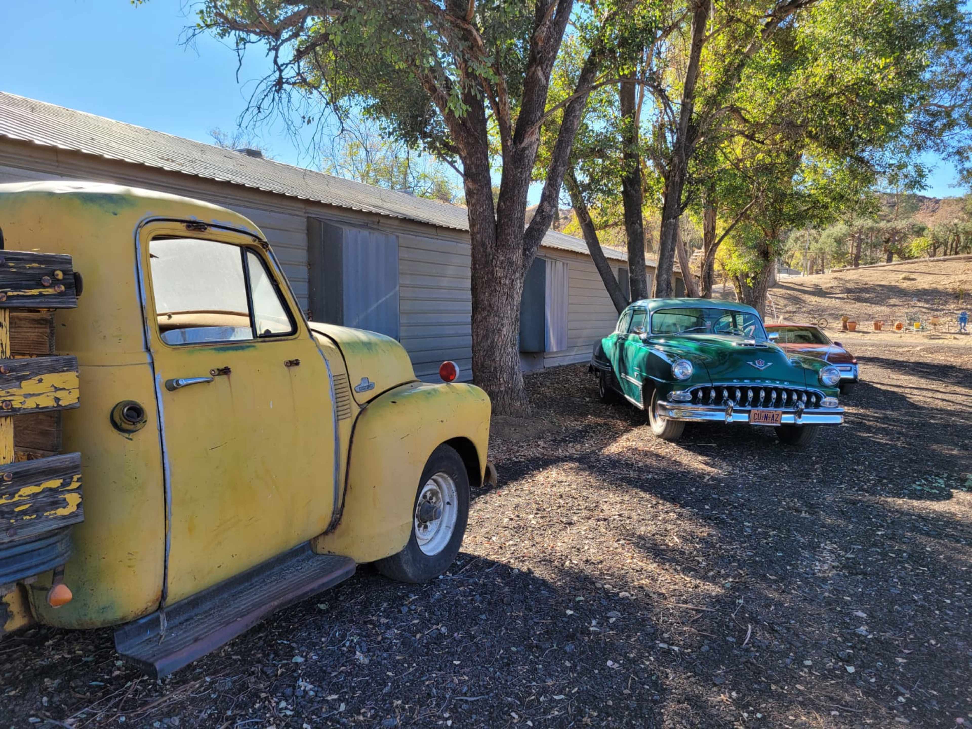 A yellow vintage truck and two classic cars parked beside a weathered building under trees.