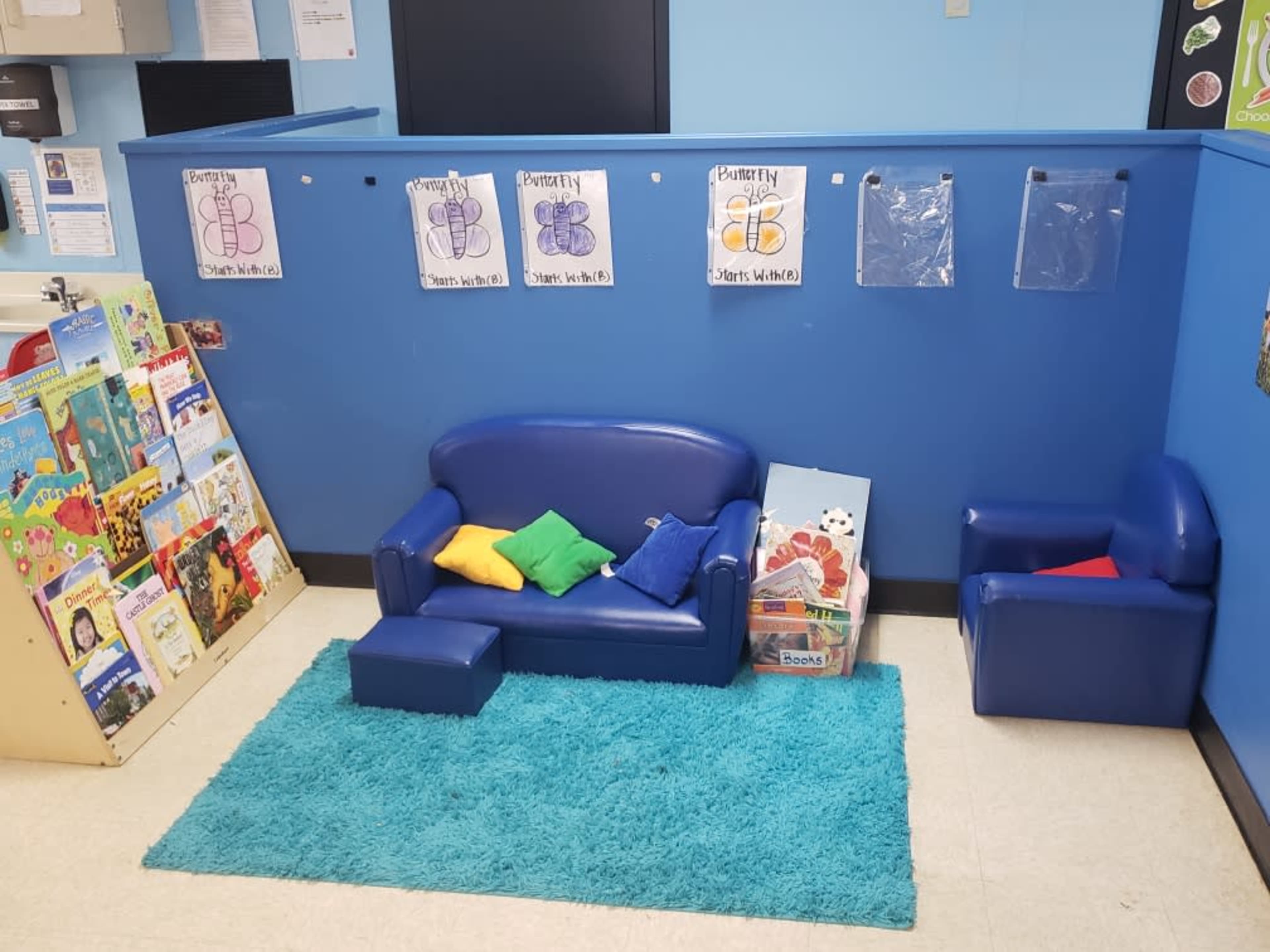 A colorful reading corner featuring a blue couch, a small armchair, a blue rug, and a low table surrounded by children's books displayed on a shelf and educational posters on the wall.