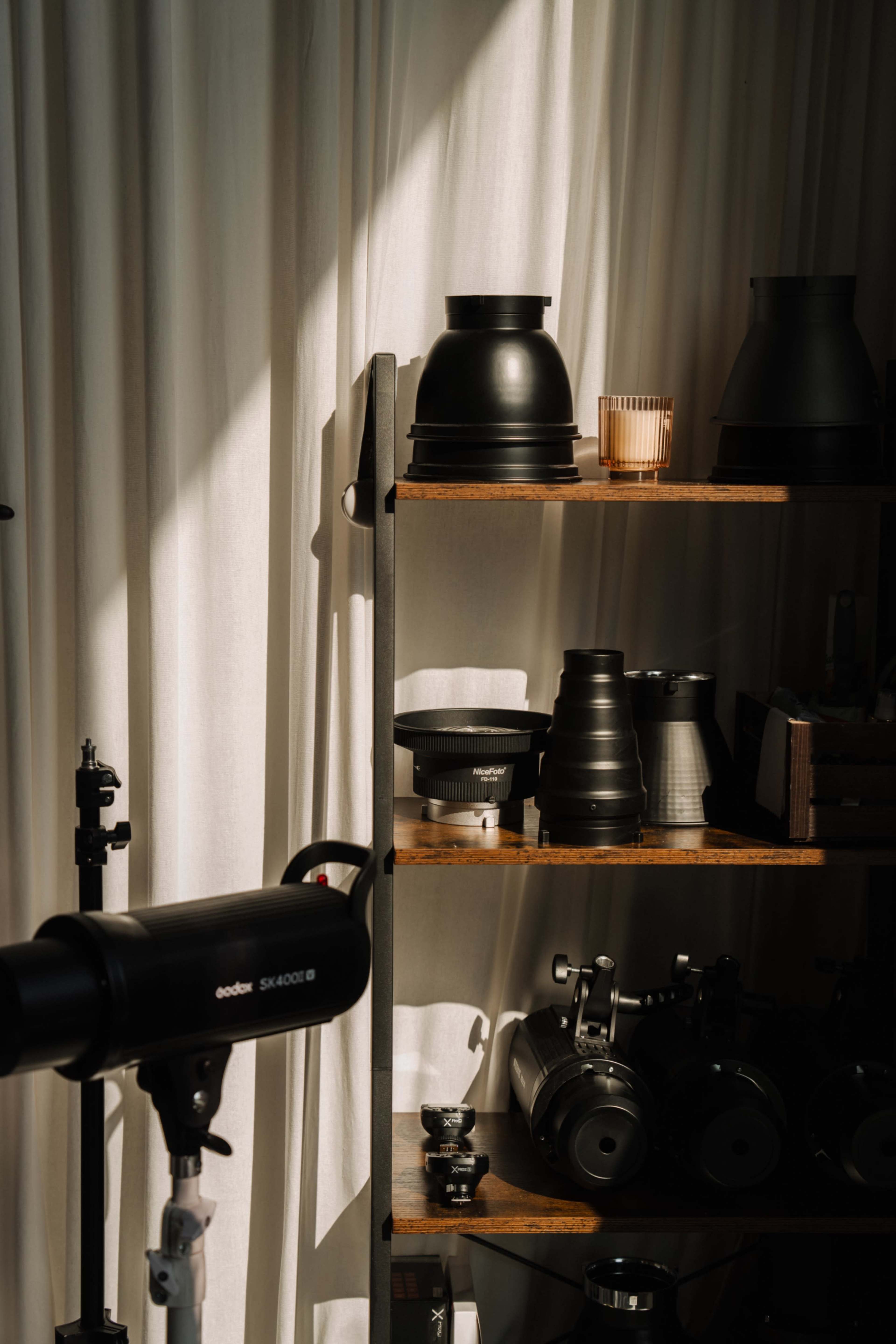 The image shows a well-organized shelf displaying various photography equipment and accessories alongside a softly lit curtain.