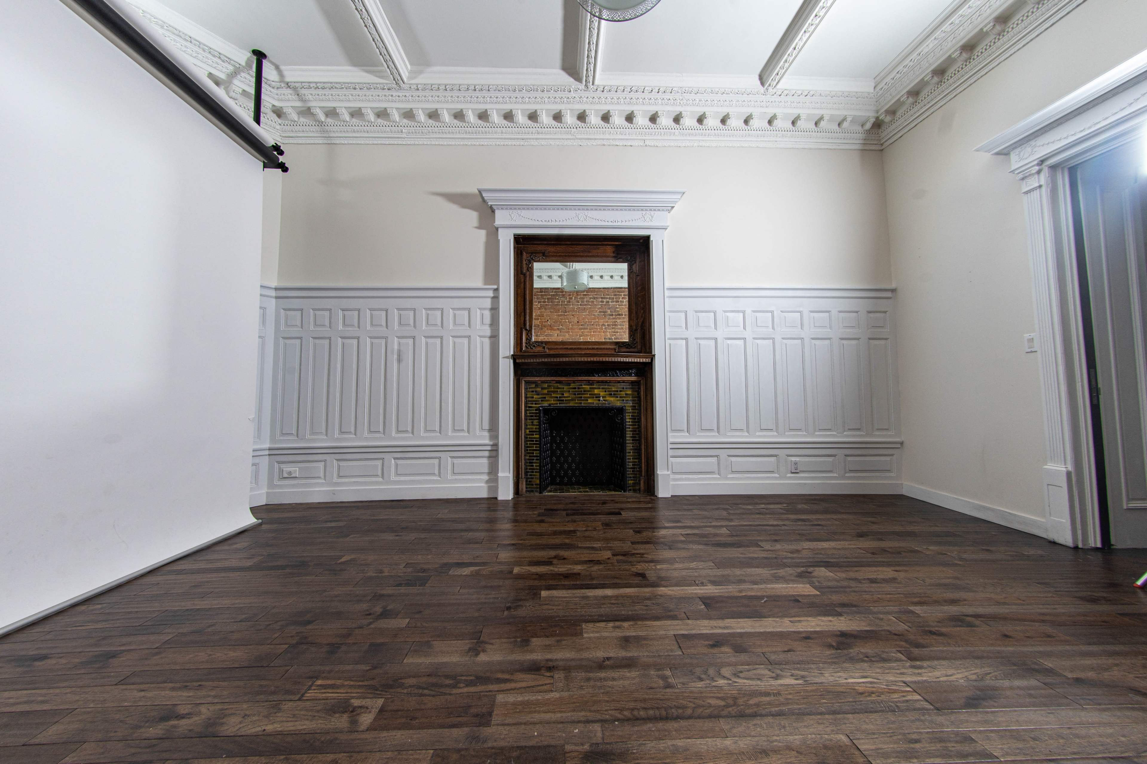 A spacious interior room with wooden flooring, wainscoted walls, and a fireplace with a mirror above it, all under a decorative ceiling.