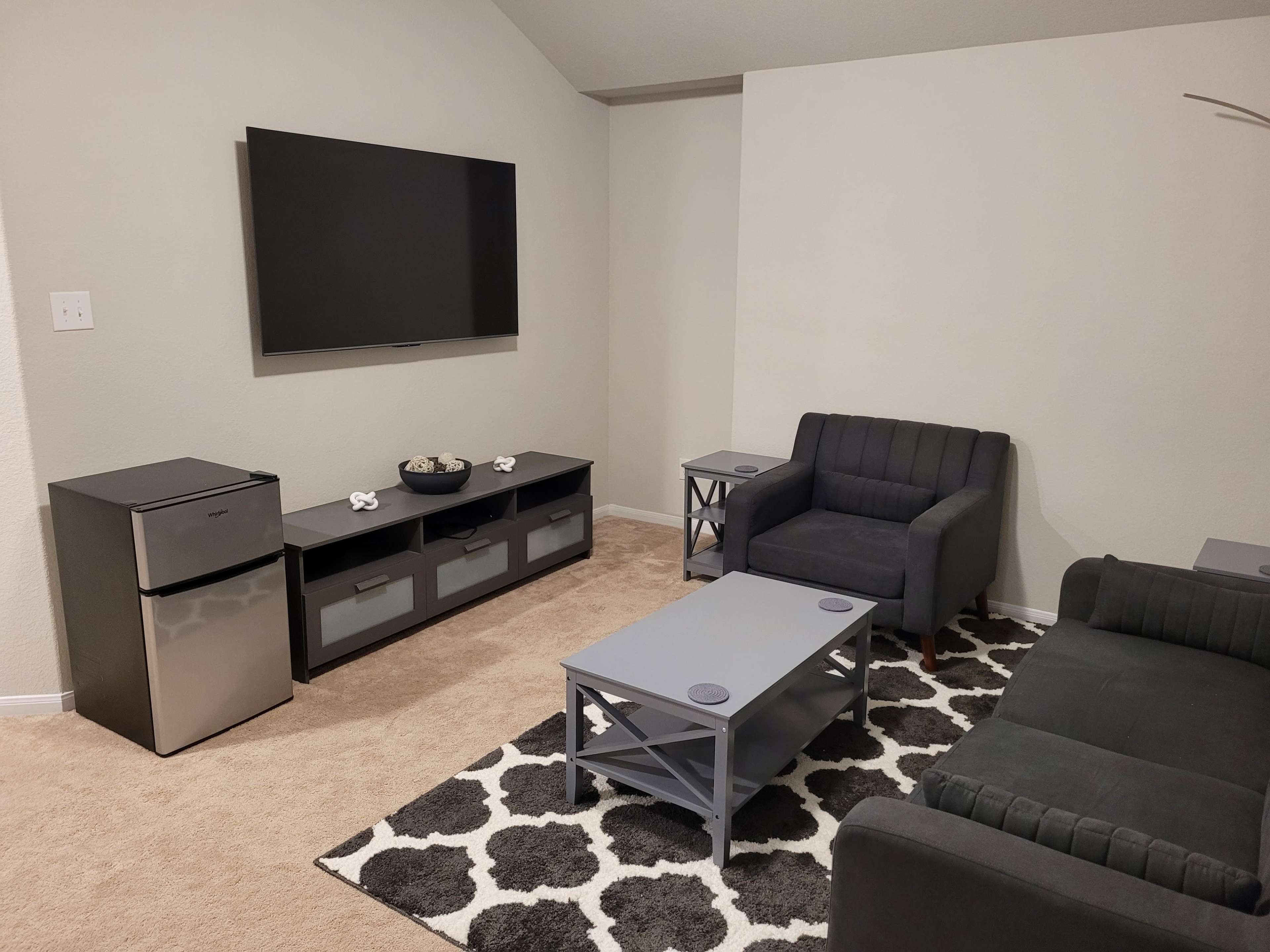 The image shows a living room with a television mounted on the wall, a gray couch, two chairs, a coffee table, an area rug, and a small refrigerator.