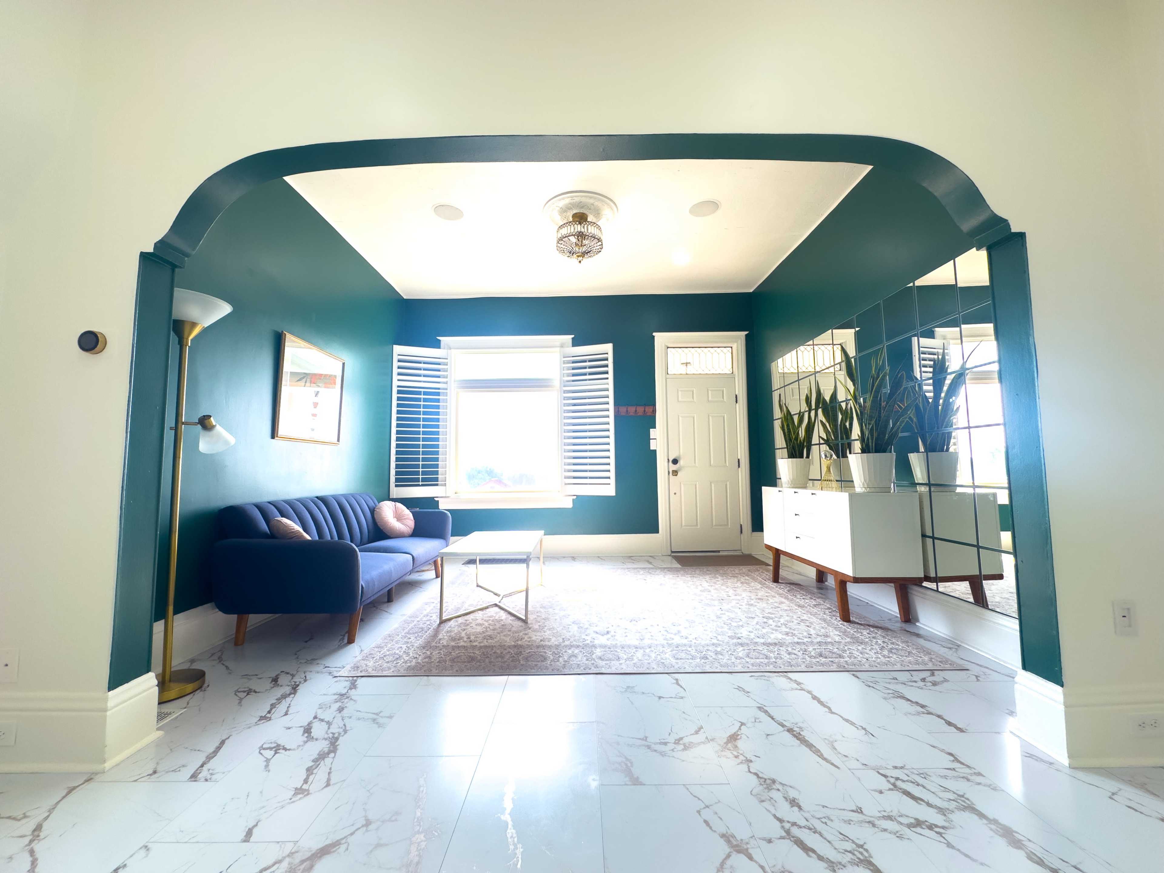 A bright living room featuring a blue sofa, a large mirror wall, and green walls with a window covered by white shutters.