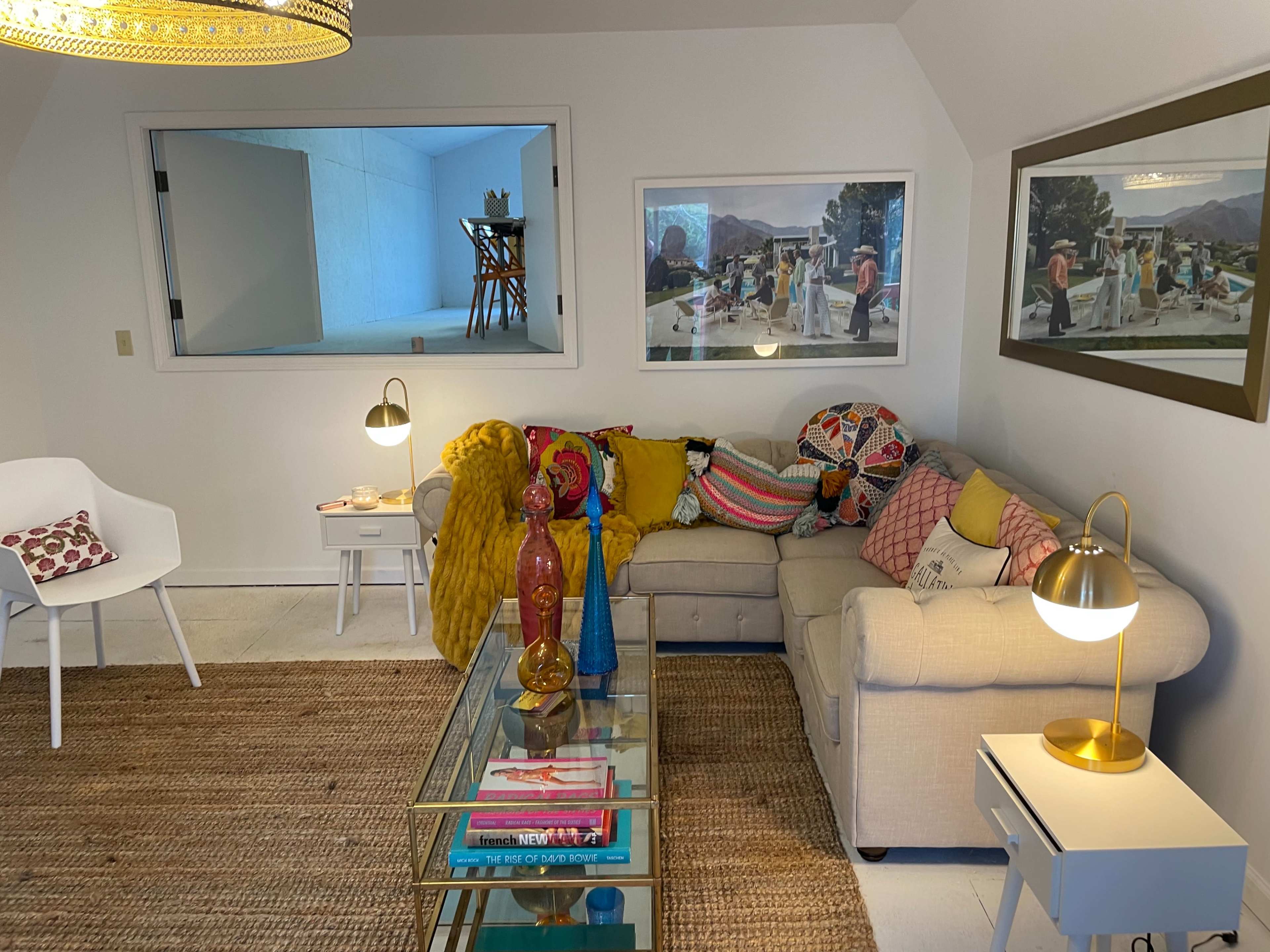 A cozy living room with a beige couch covered in colorful pillows, a glass coffee table, and a chair, with framed pictures on the wall and a large window revealing an adjacent room.