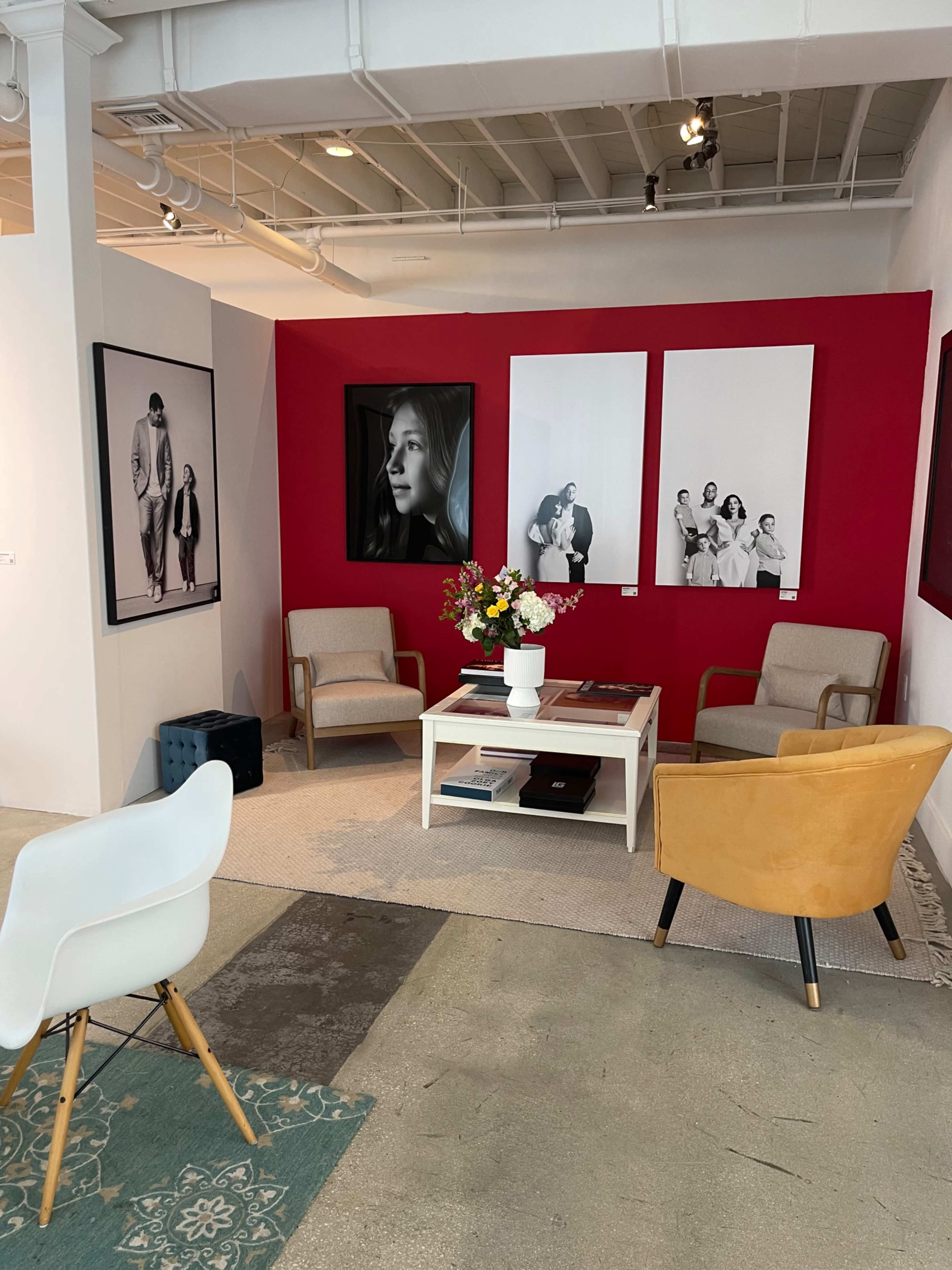 The image showcases a modern art gallery with a seating area featuring two armchairs, a coffee table, and framed black-and-white photographs on a red accent wall.