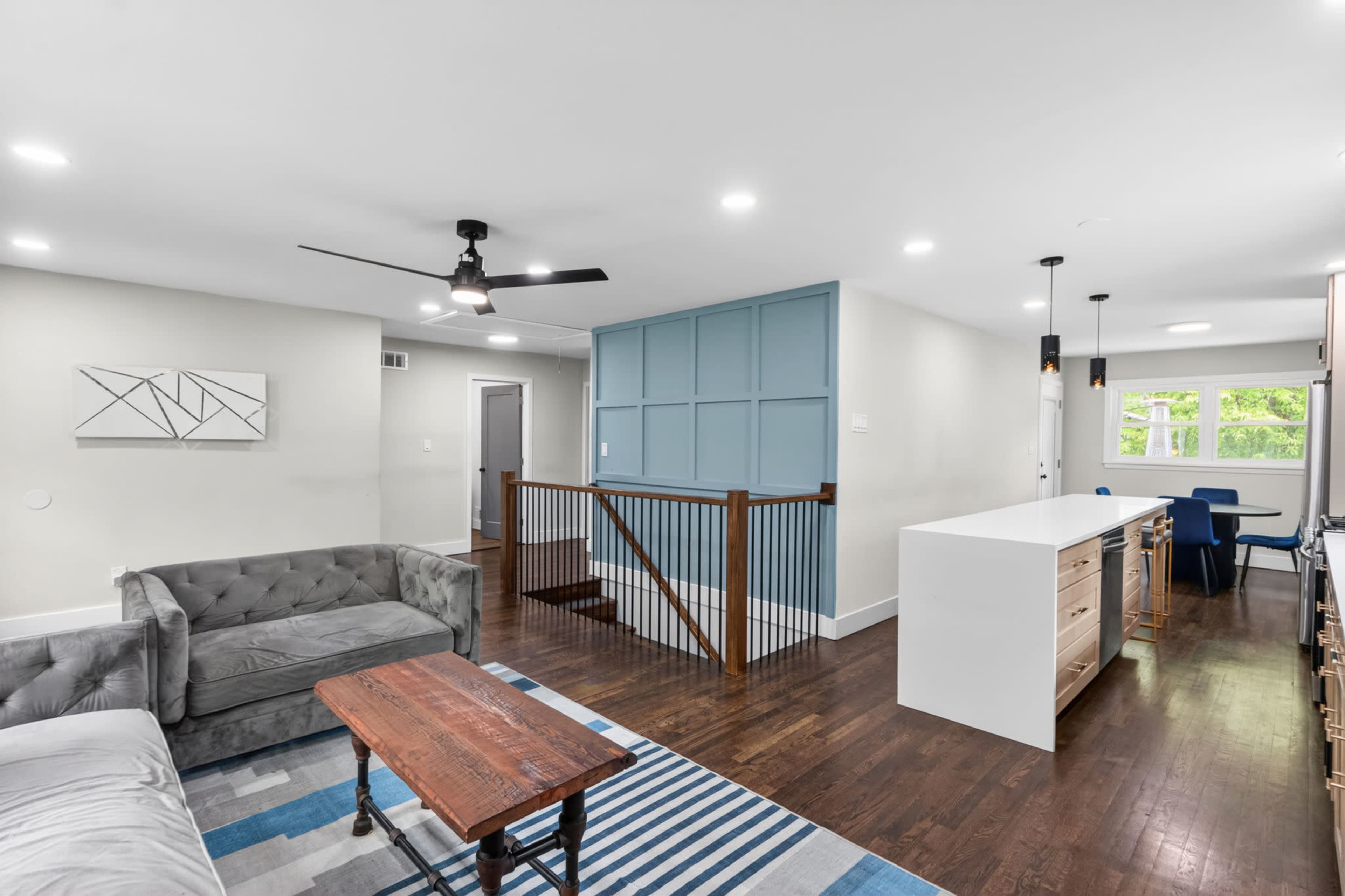 The image shows a modern, open-concept living space featuring a gray sofa, a wooden coffee table, and a white kitchen island with a blue accent wall in the background.