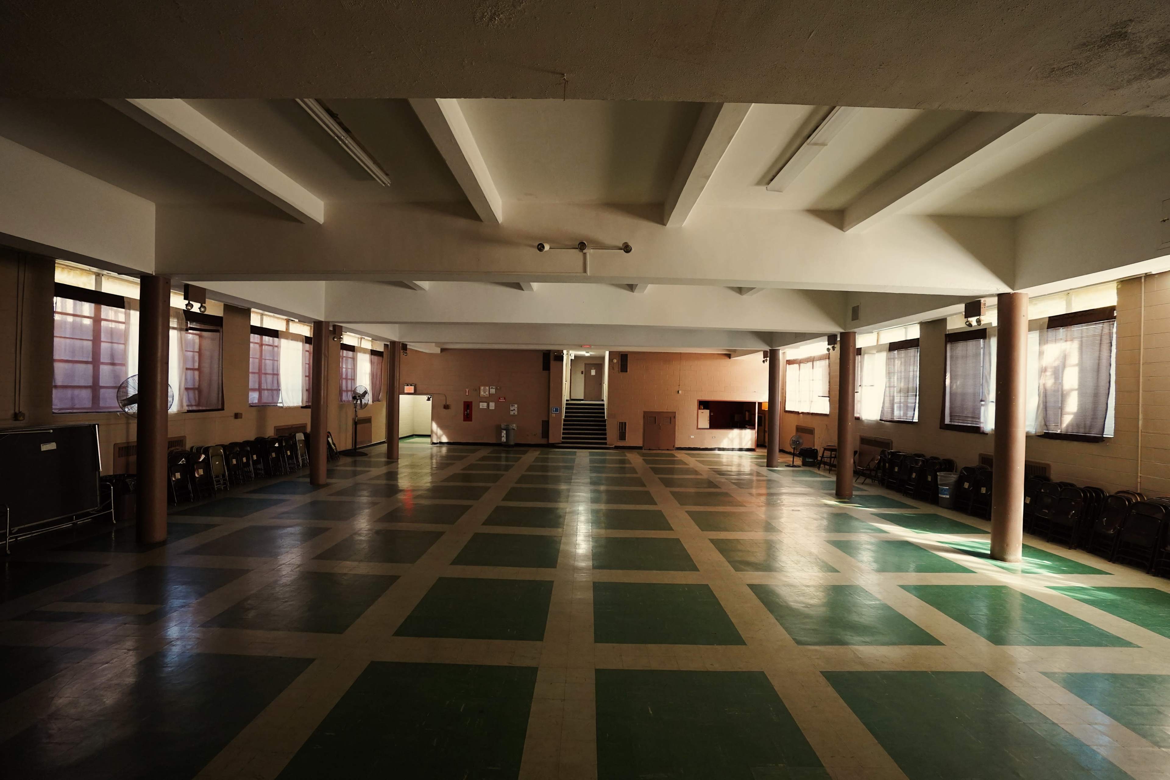 A large, empty hall with green and beige tiled flooring and sunlight streaming through the windows.
