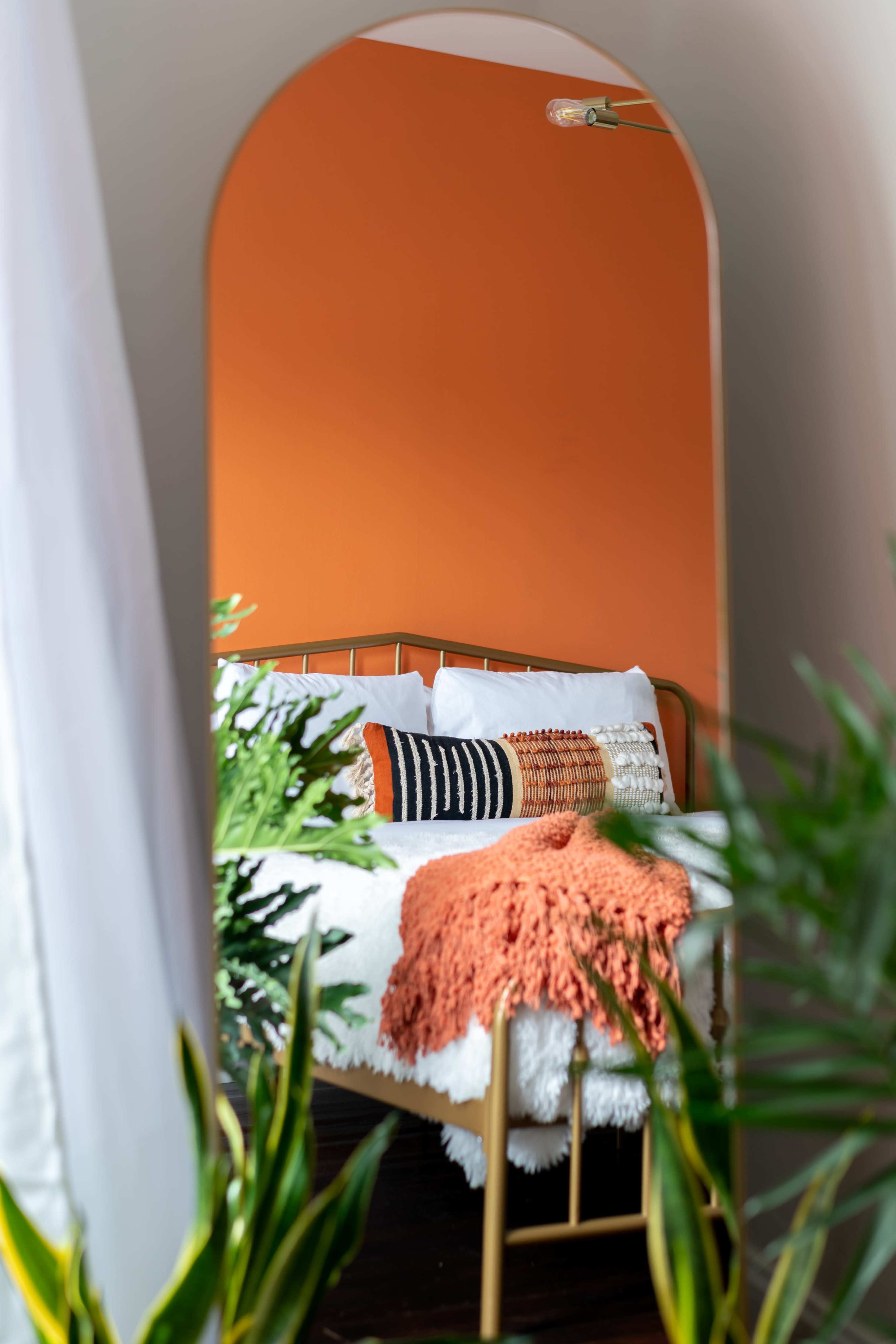A mirror reflects a bedroom featuring an orange wall, a metal bed with decorative pillows, and green plants surrounding the scene.