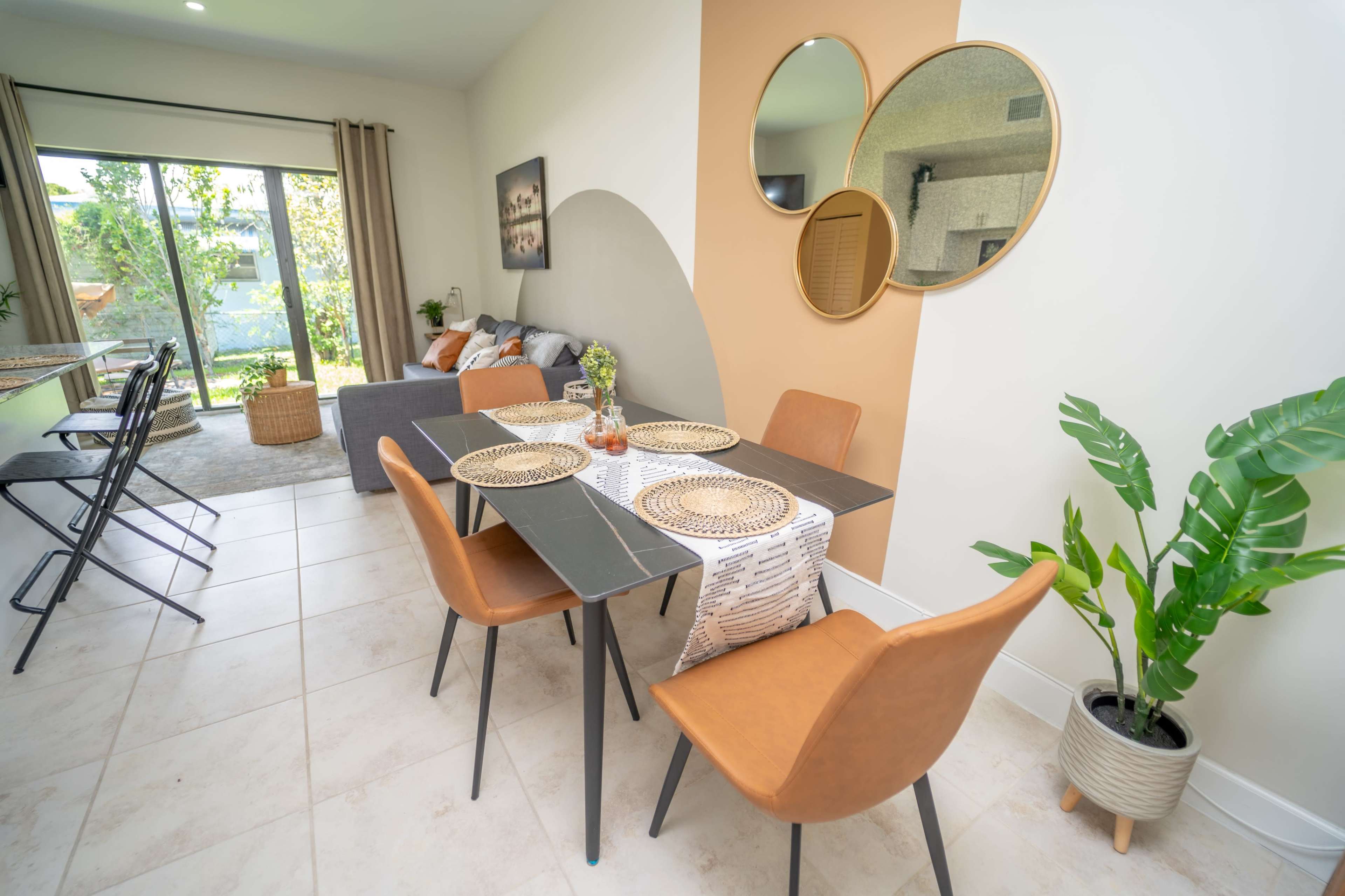 A modern dining area features a table set for four, surrounded by brown chairs, with a living room and kitchen visible in the background.