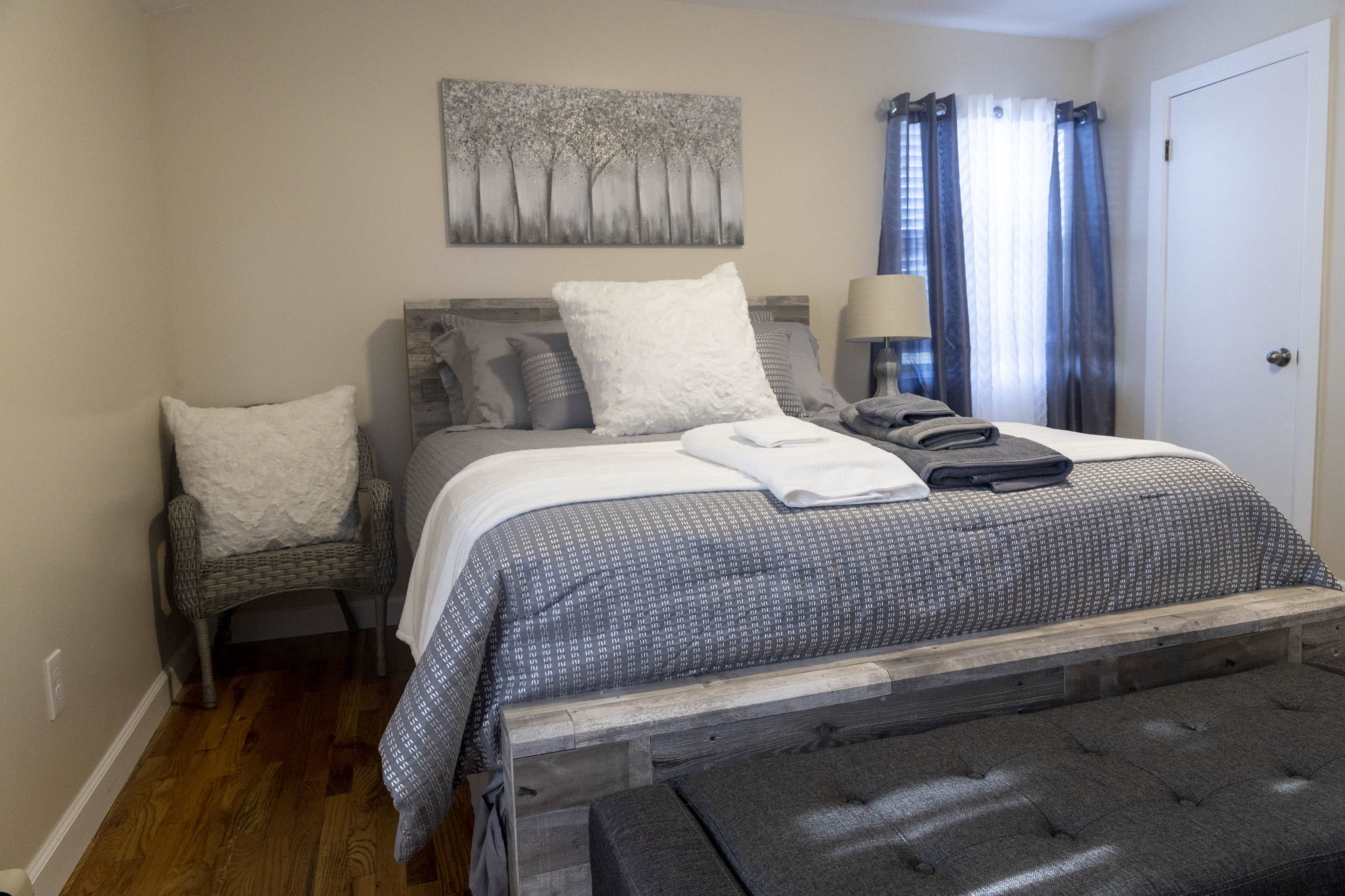 A neatly arranged bedroom with a wooden bed, gray bedding, a decorative wall art of trees, and a chair in the corner.