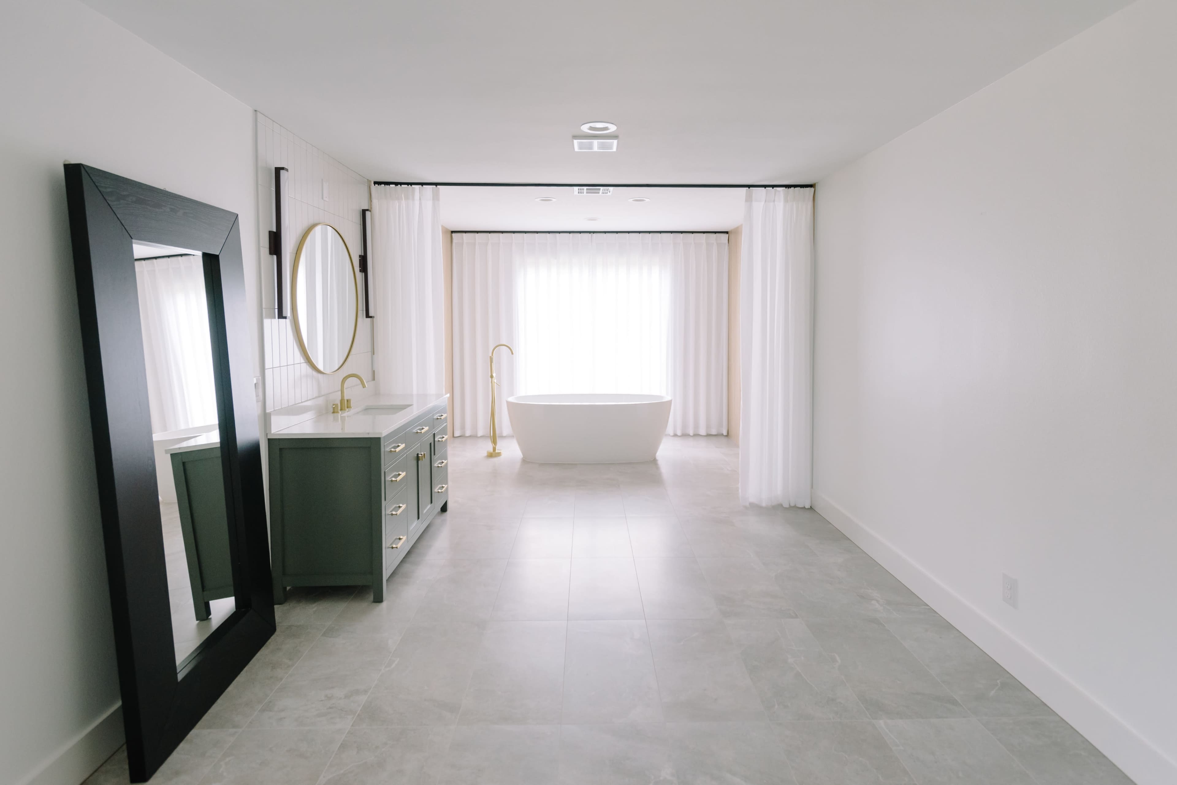 The image shows a minimalist bathroom featuring a freestanding bathtub, a sink area with a large mirror, and natural light streaming through sheer curtains.