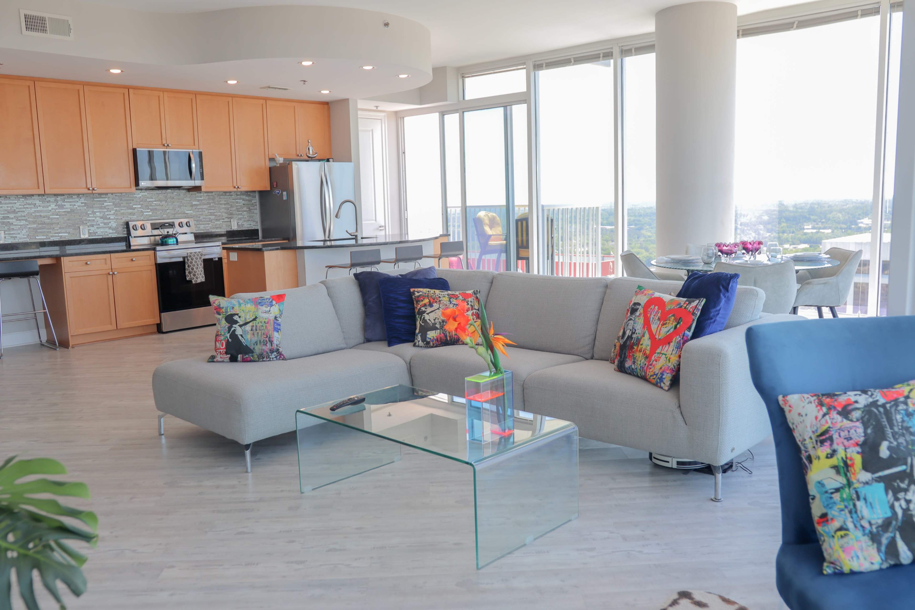 A modern, open-concept living area with a sectional sofa, glass coffee table, and a kitchen in the background featuring wooden cabinets and stainless steel appliances.