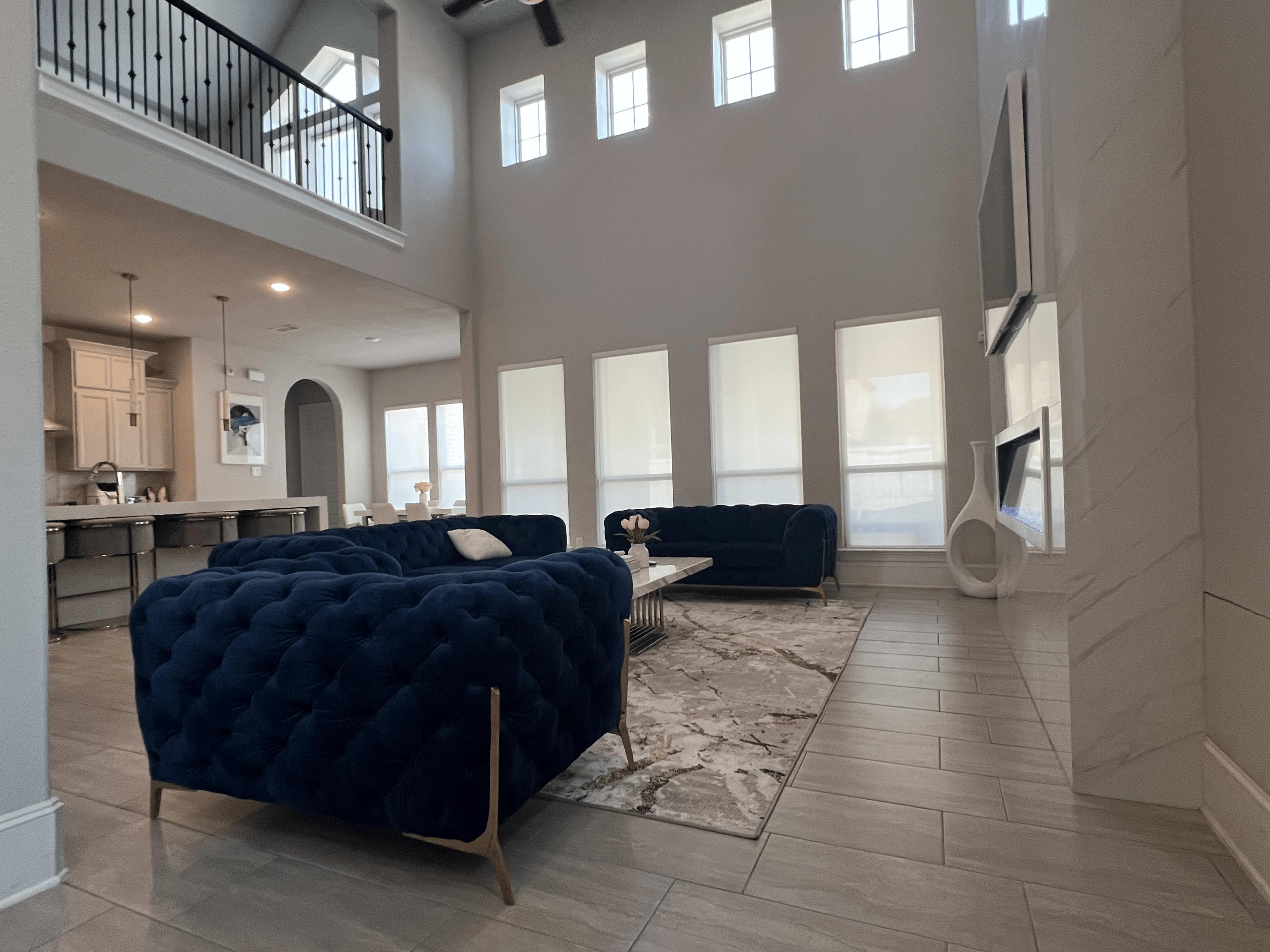 The image shows a spacious living room with two blue tufted sofas facing a large, light-colored area rug, and a modern kitchen visible in the background.