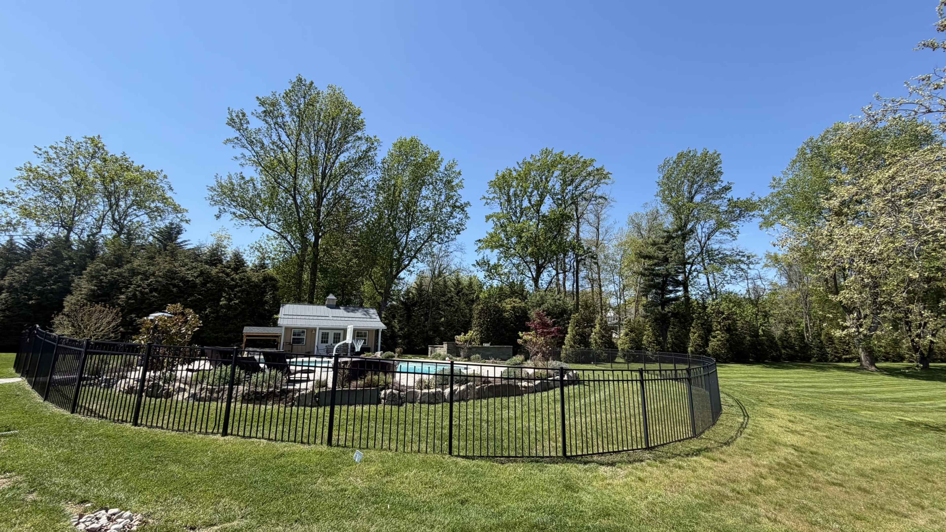 A fenced backyard features a small shed, a swimming pool, and numerous trees against a clear blue sky.
