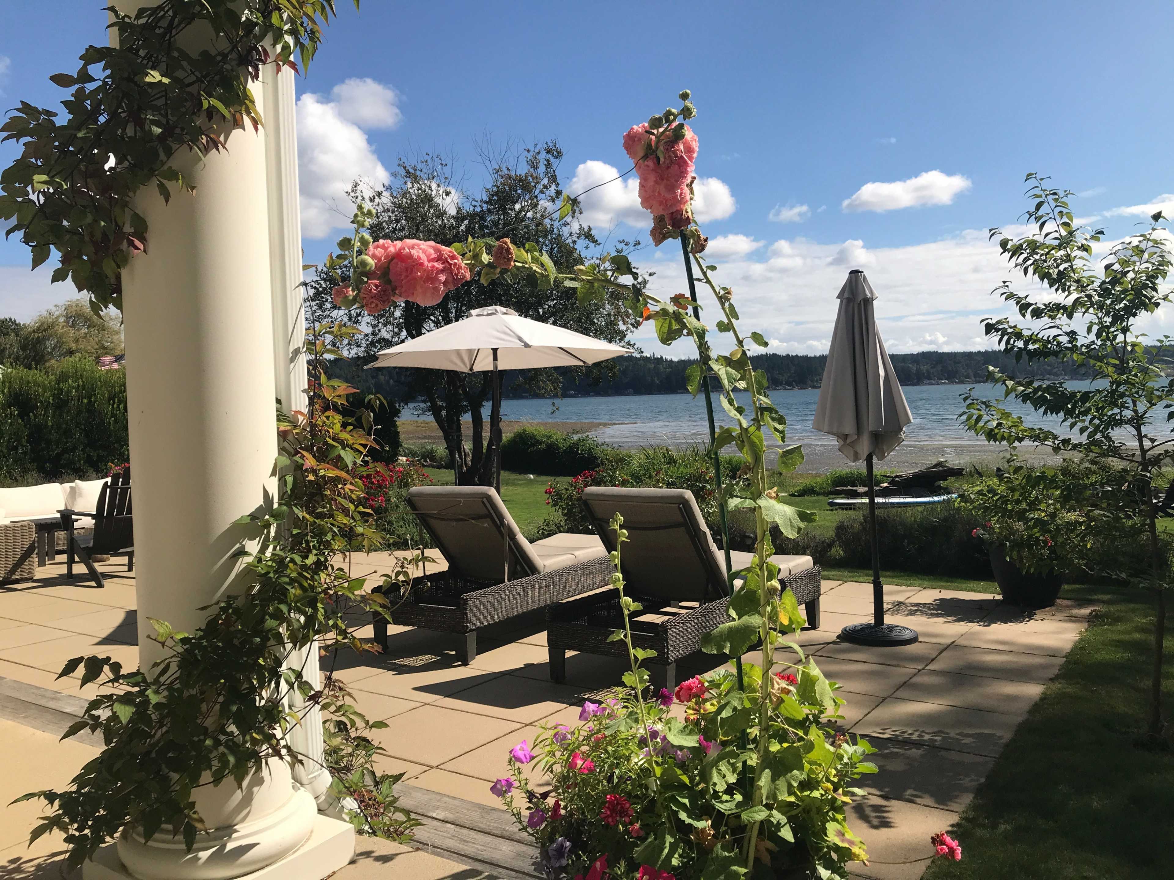 A patio area features two lounge chairs beside a lake, with flowers and umbrellas framing the view.