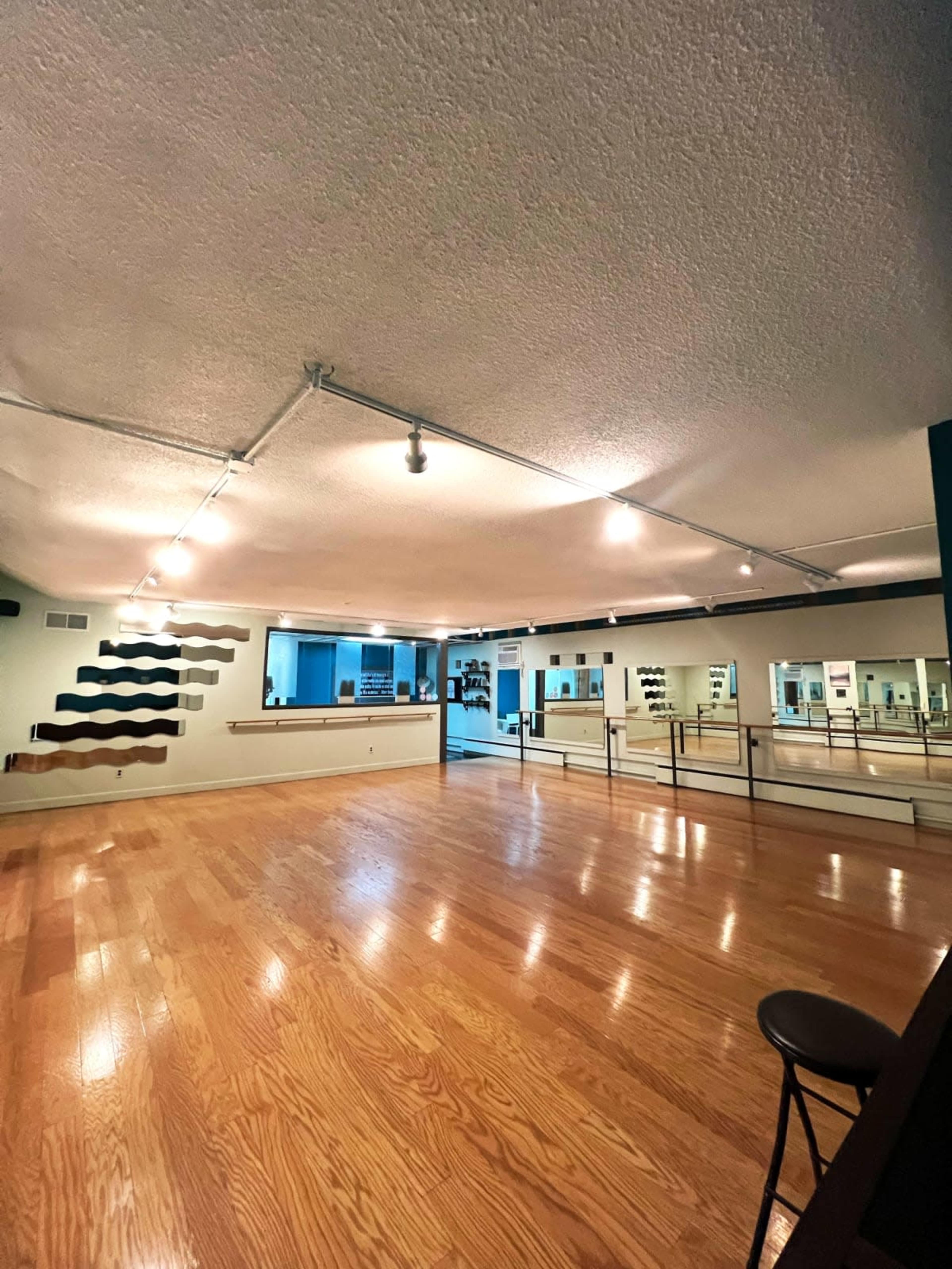 The image shows a spacious studio with wooden floors, mirror walls, and track lighting, featuring a staircase along one side.