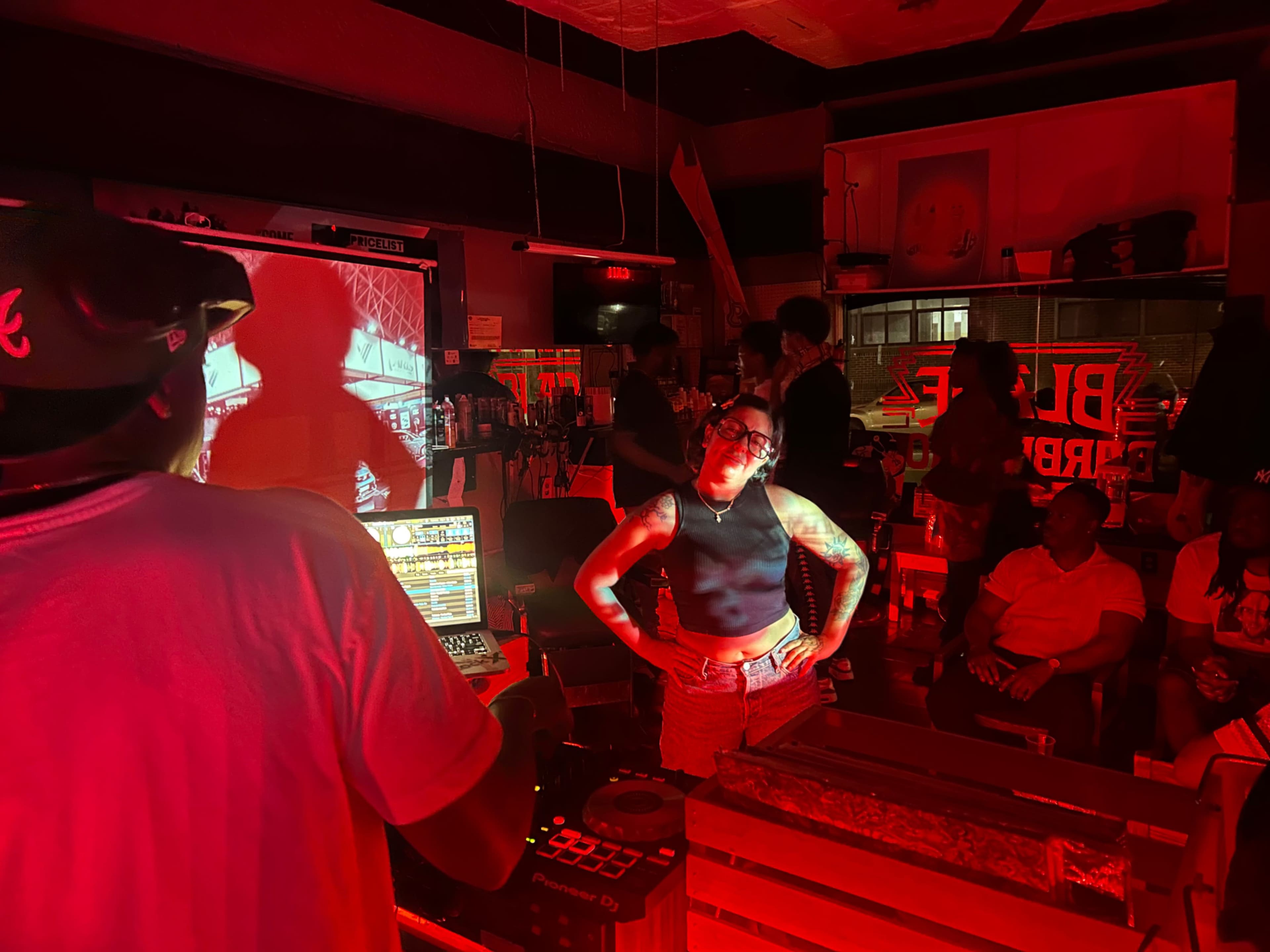 A DJ stands behind a laptop in a dimly lit room filled with people socializing and dancing, illuminated by red lighting and a projection on the wall.