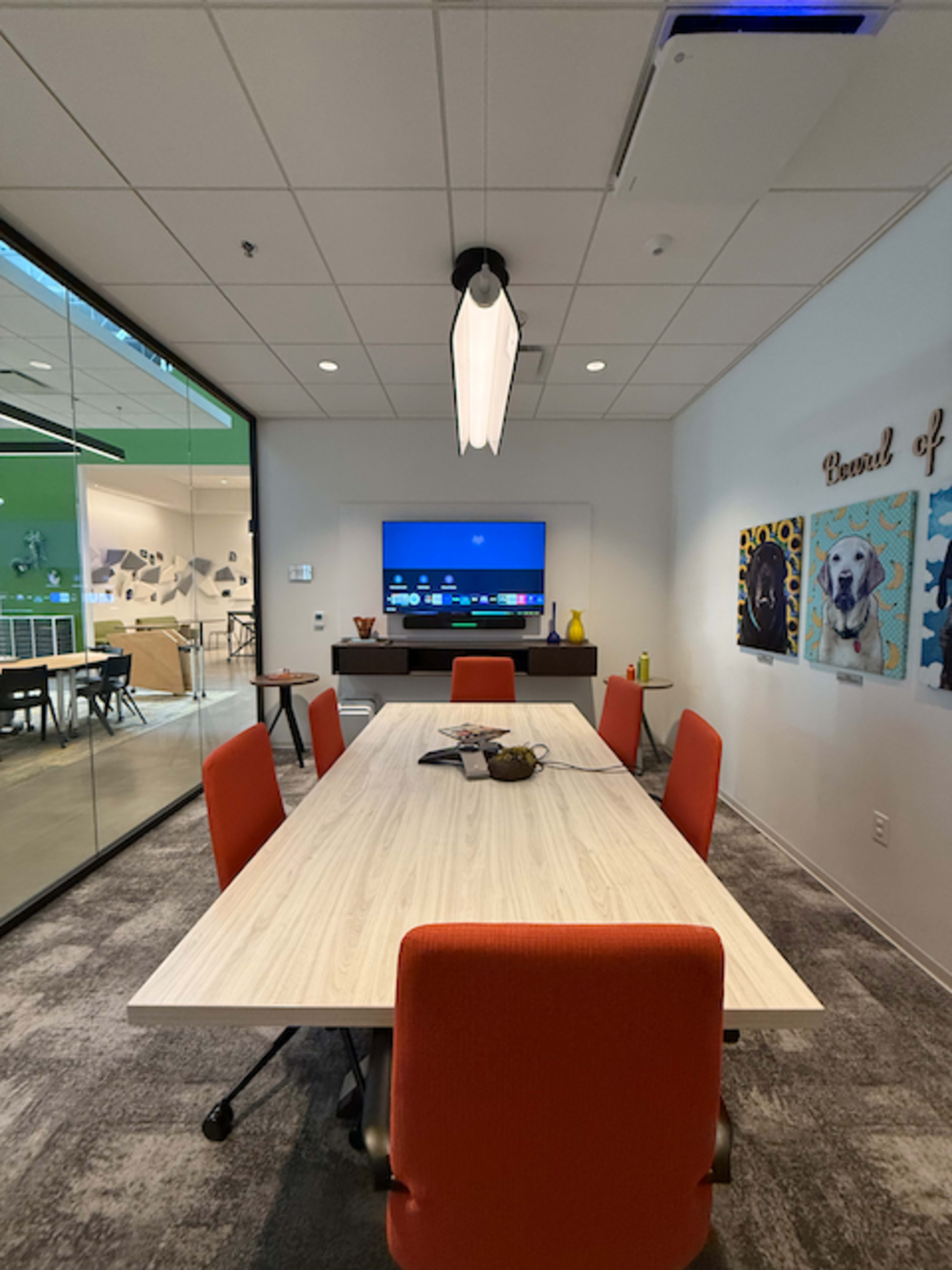 The image shows a modern conference room with a long table surrounded by orange chairs, featuring a television mounted on the wall and artwork depicting dogs.