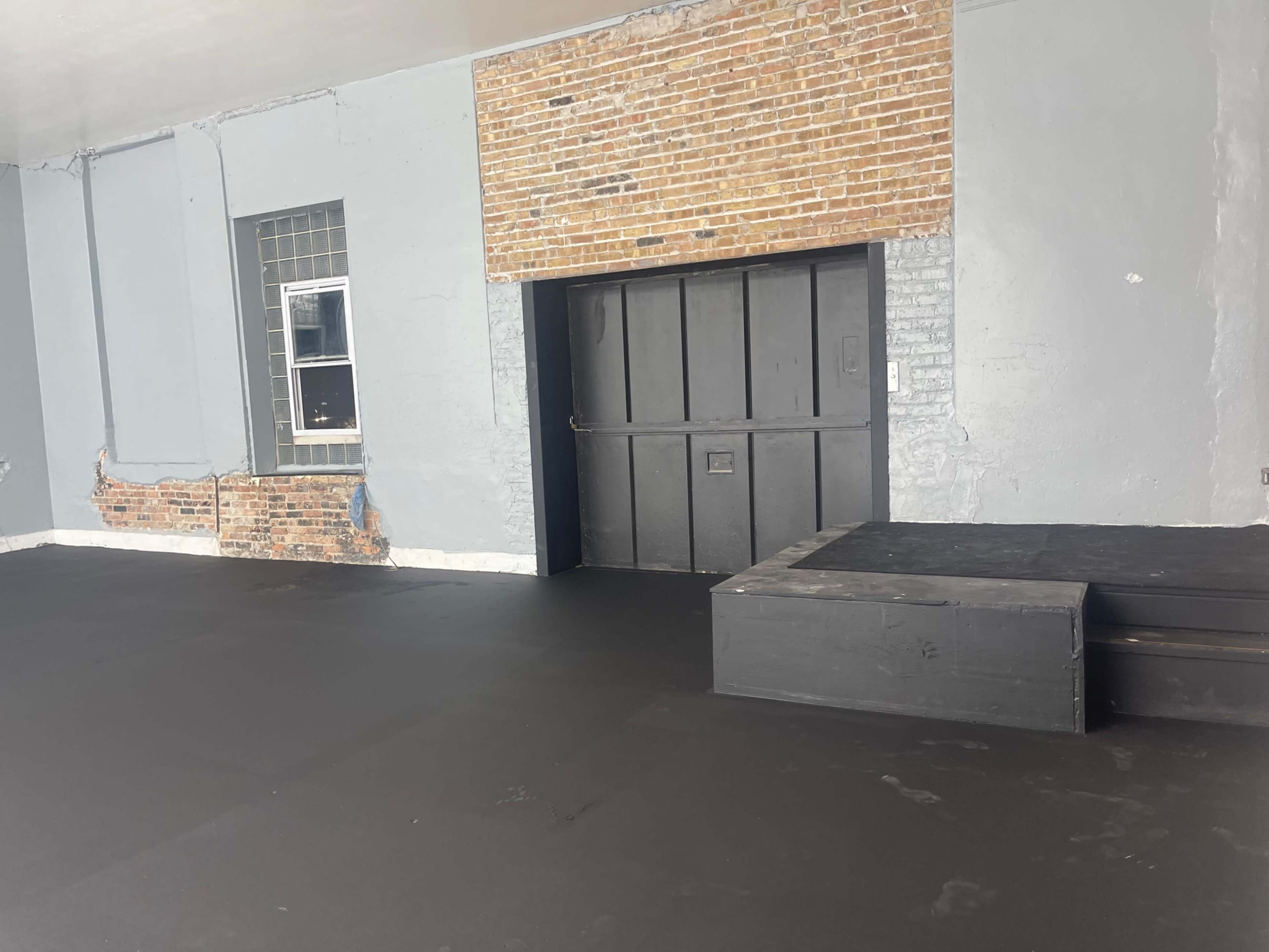 An empty room with a gray wall, a brick section, a closed door, and black flooring.