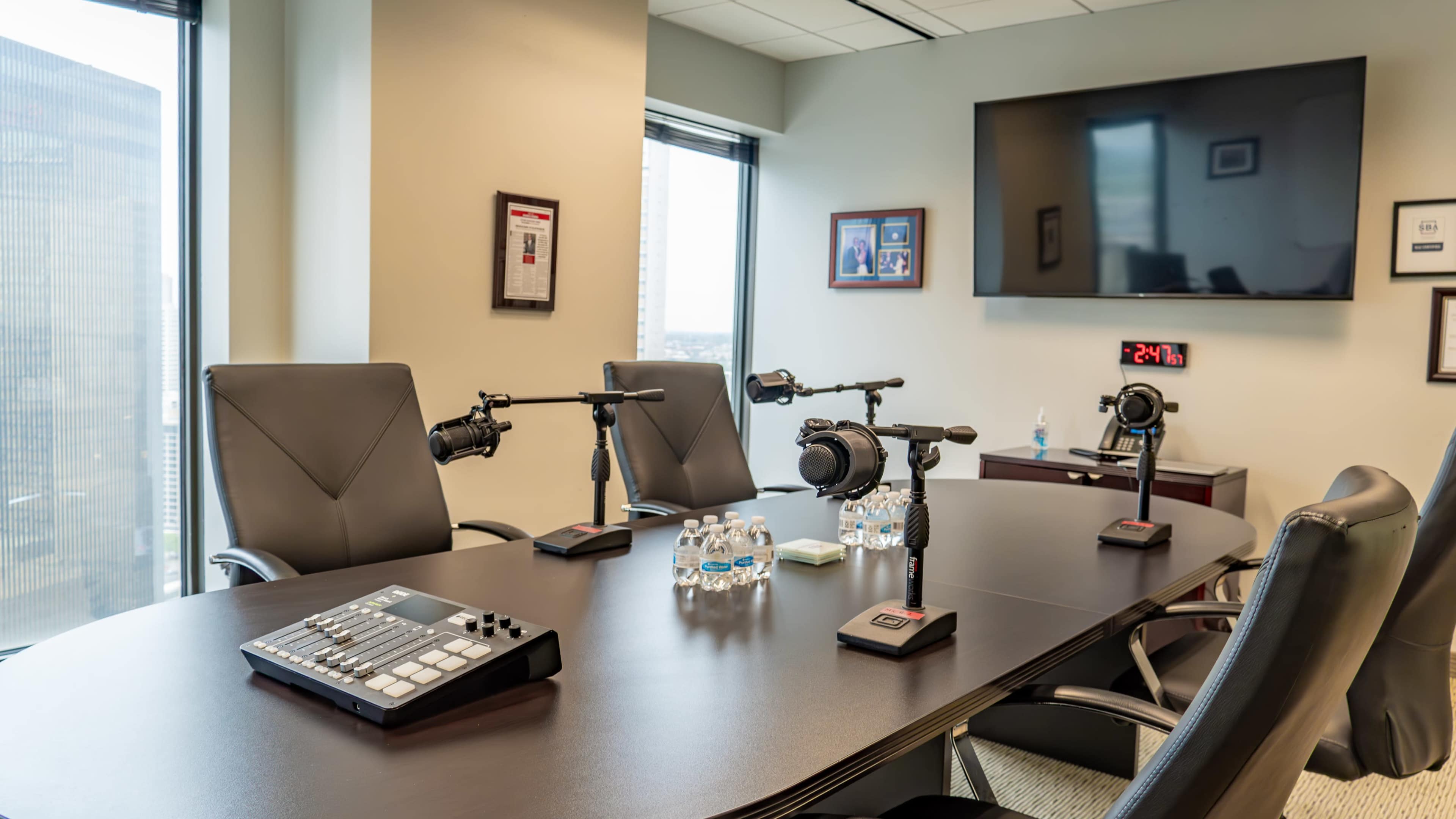 A conference room is set up with a large table, three microphones mounted on stands, a digital mixing board, and water bottles, with windows displaying a cityscape in the background.