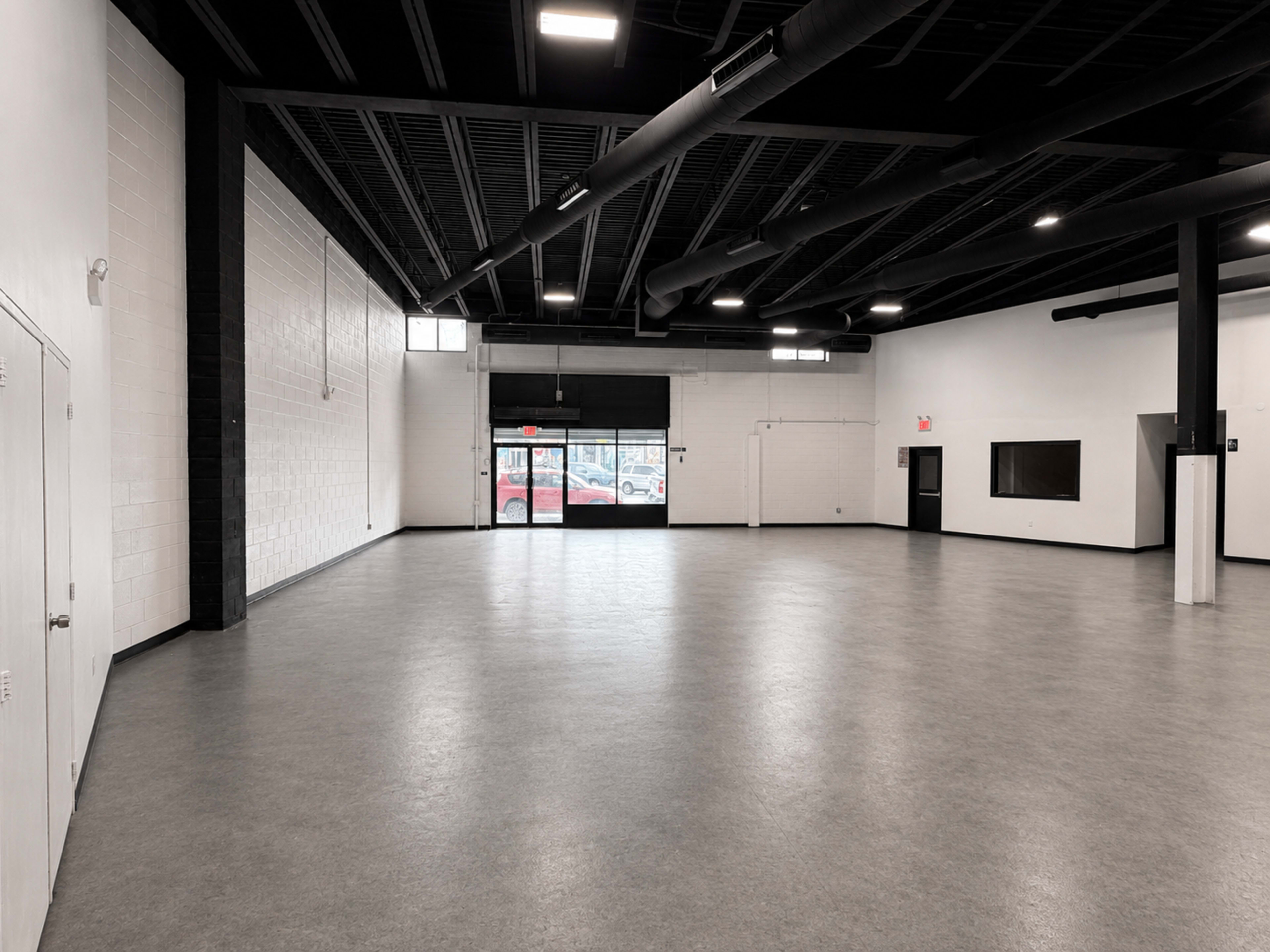 The image shows a large, empty room with gray flooring, black beams overhead, and a doorway leading outside.