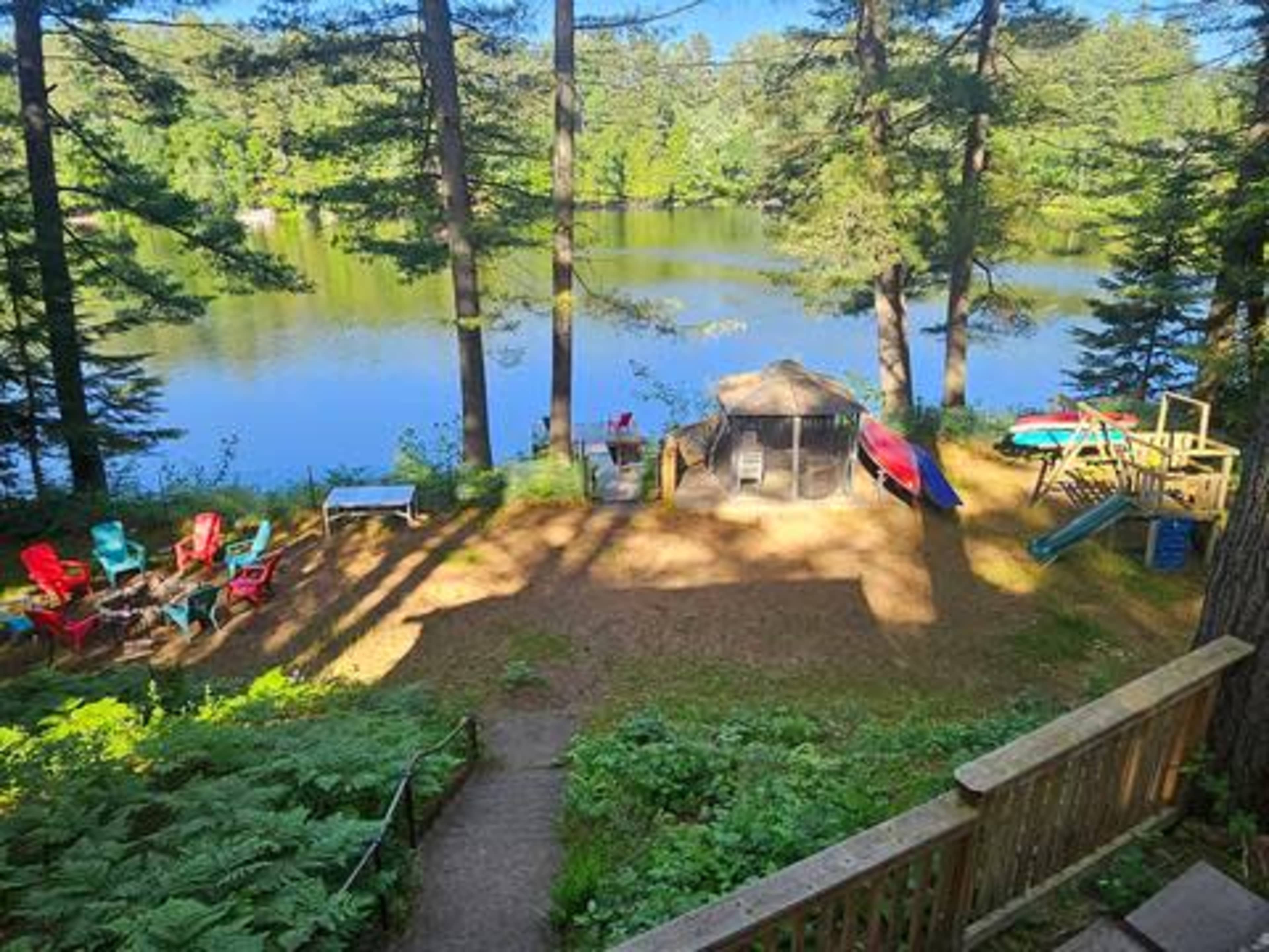 The image shows a lakeside campsite featuring various sitting areas, a large tent, and a playground, surrounded by tall trees.