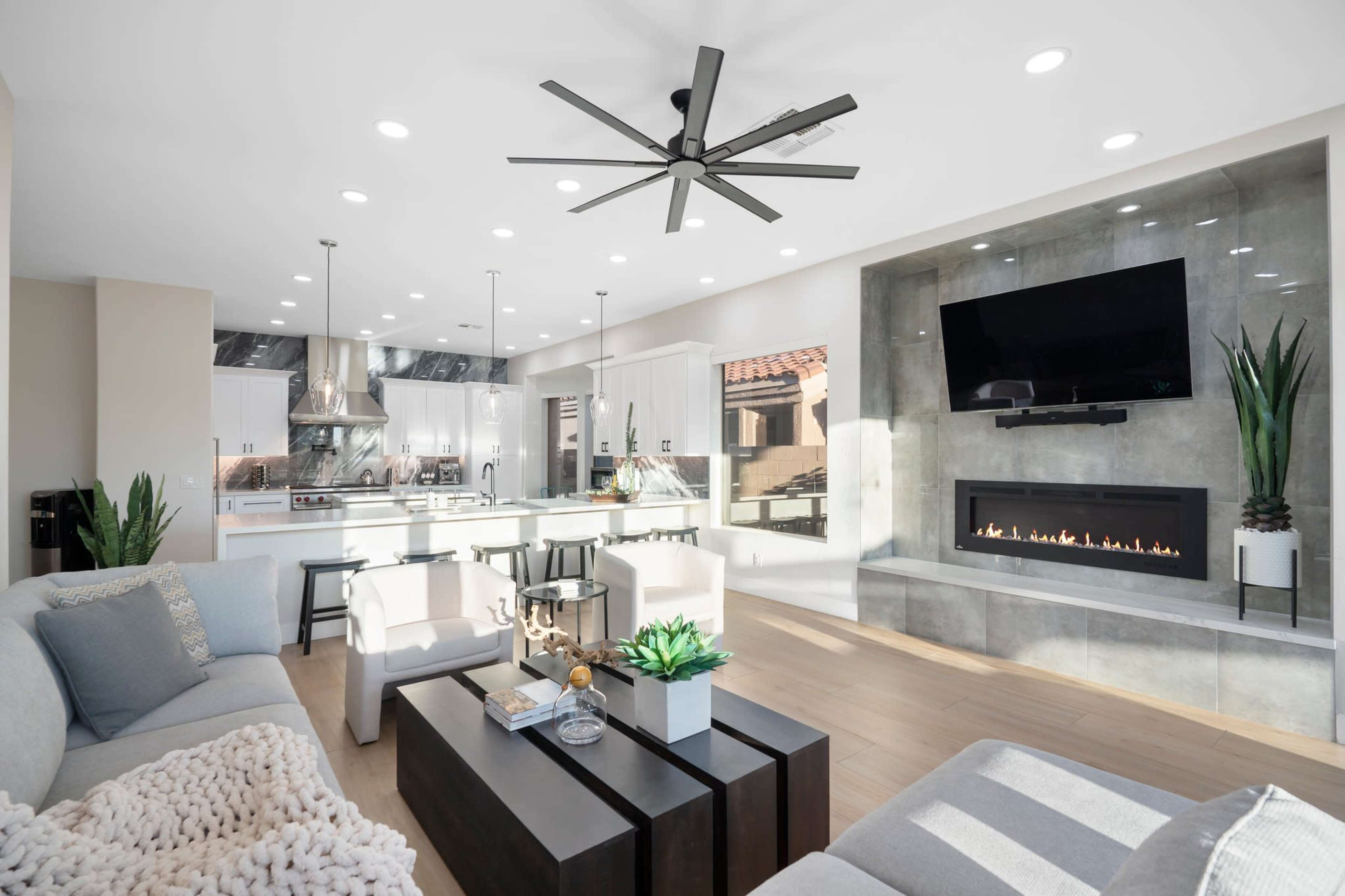 A modern living room and kitchen area featuring a large sectional sofa, a coffee table, a flat-screen TV mounted on the wall, and a fireplace.