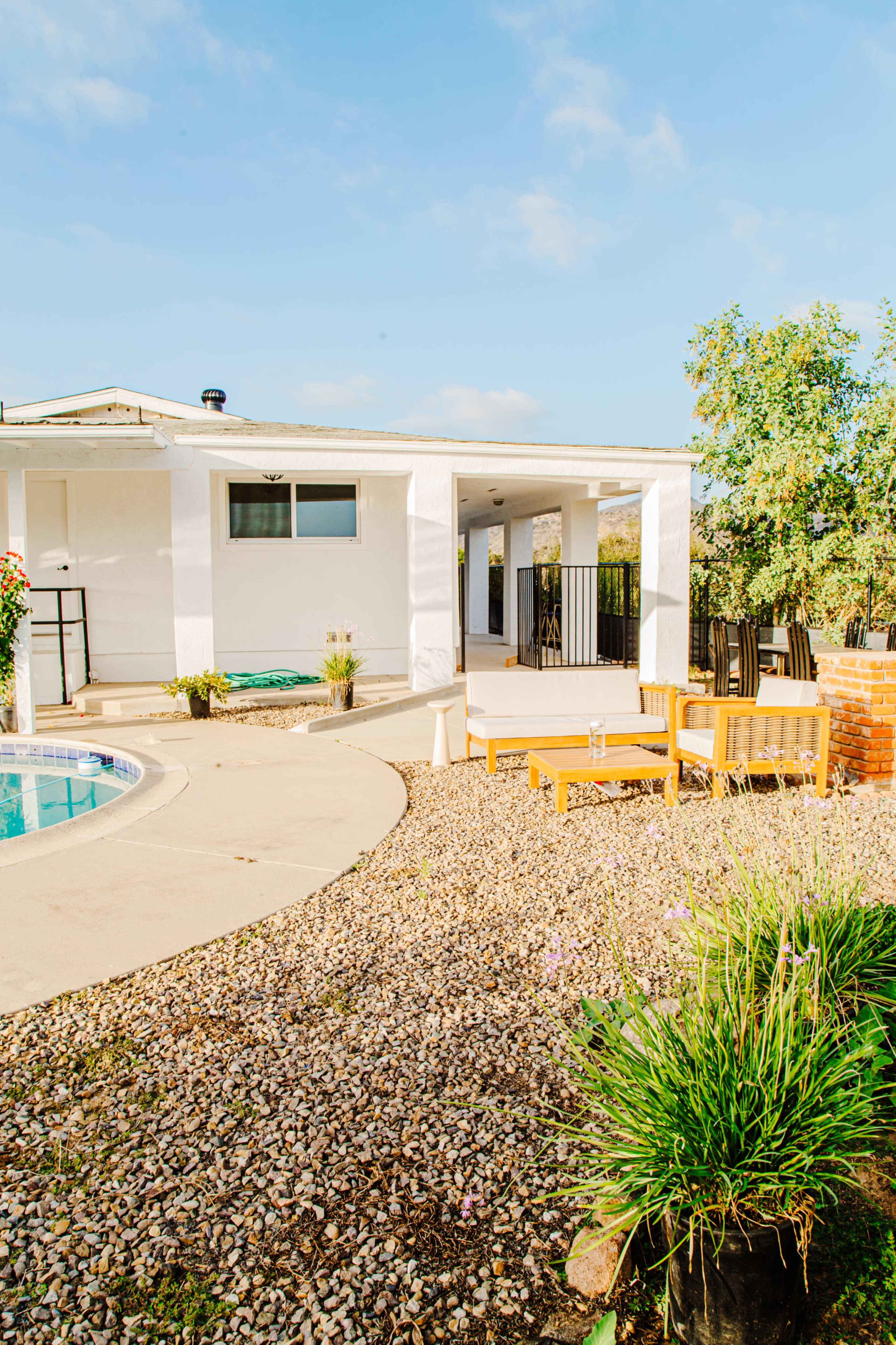 A backyard features a swimming pool, gravel landscaping, wooden seating areas, and a white house with a covered porch.