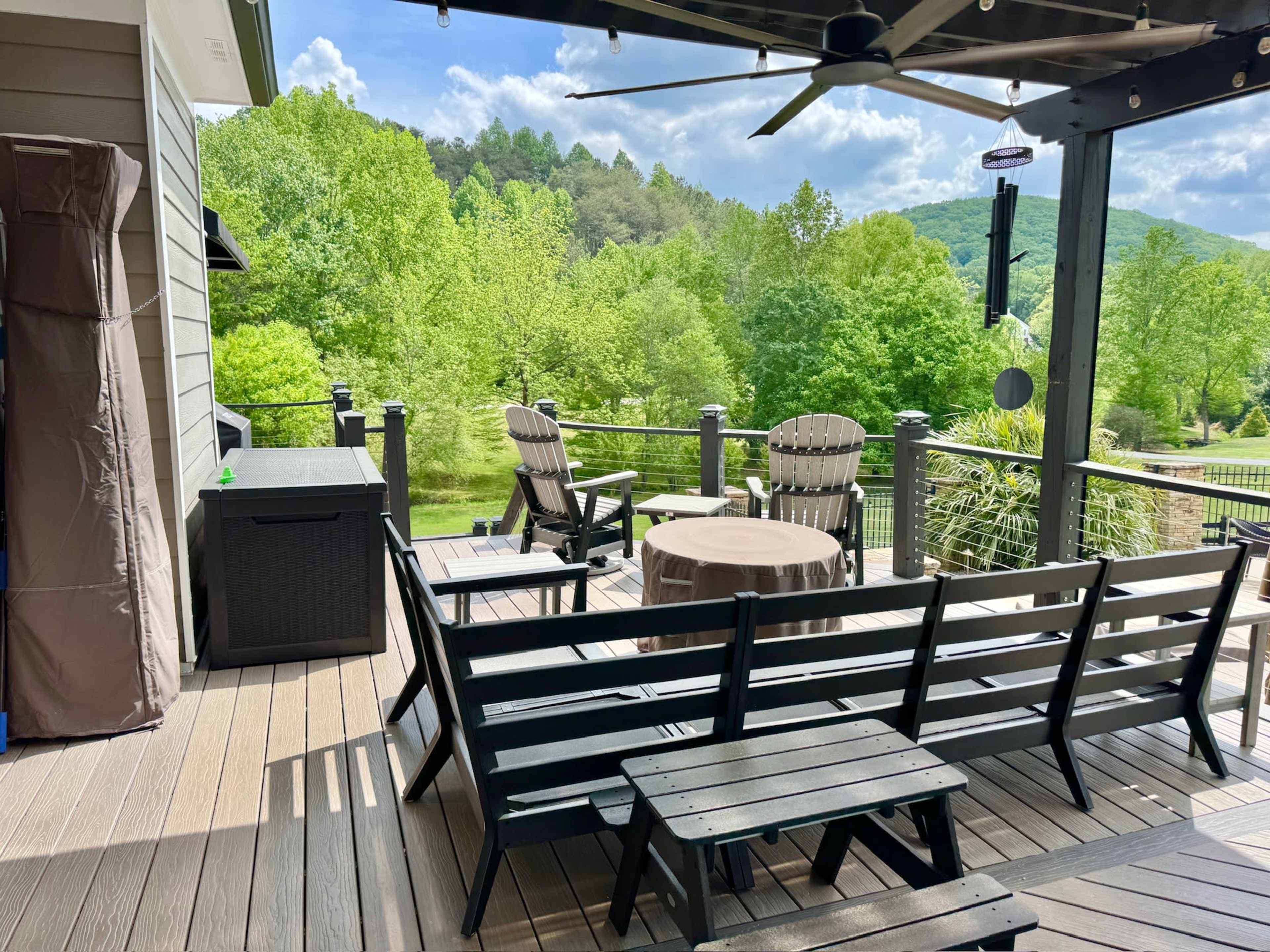 A deck with black wooden furniture overlooks a green landscape of trees and hills under a partly cloudy sky.