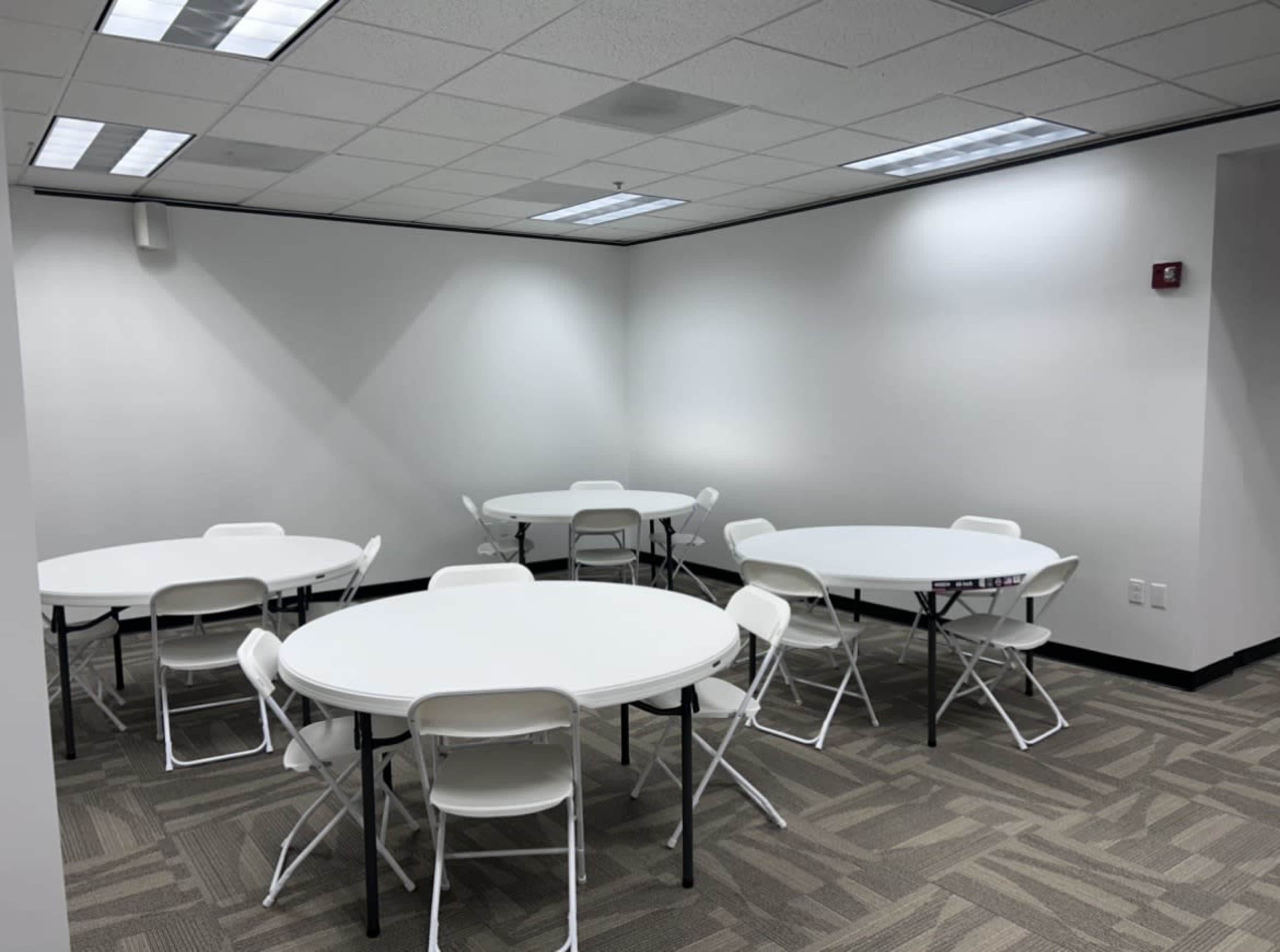 The image shows a room with several round tables and white folding chairs arranged neatly on a patterned carpet.