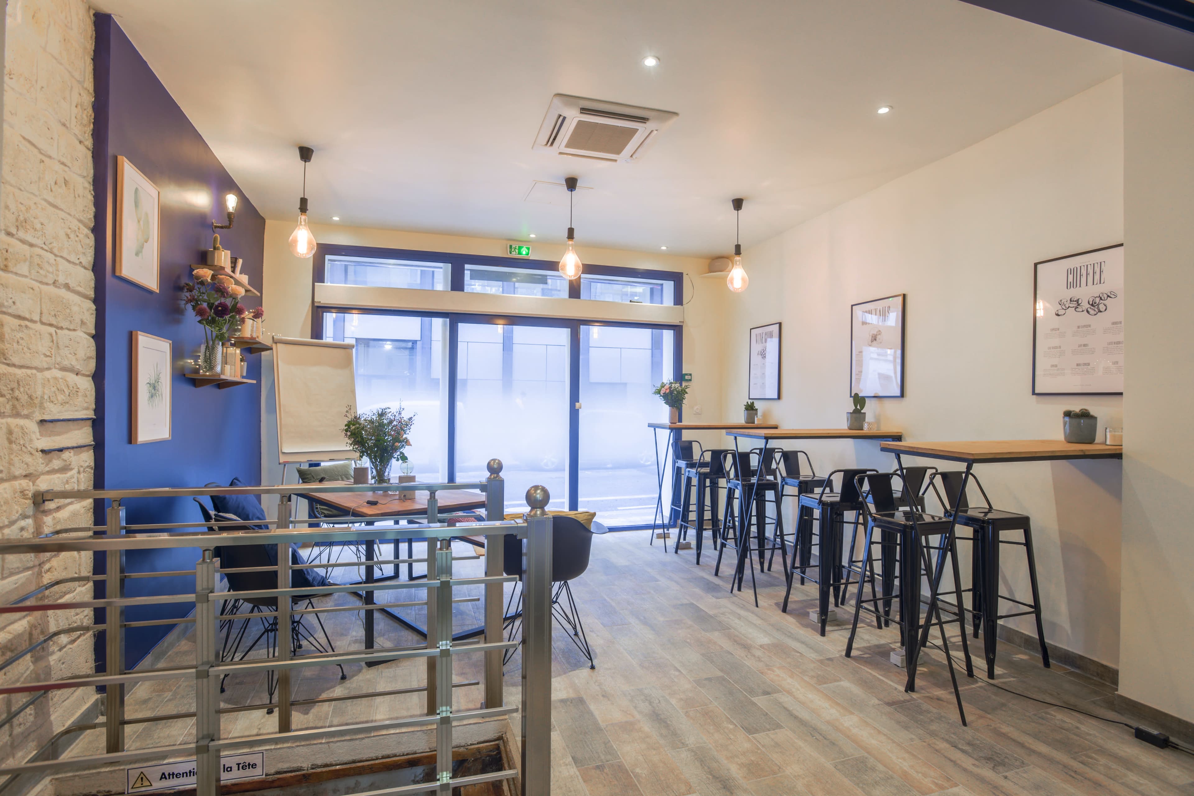 The interior of a café features a mix of high tables and chairs along one wall, with decorative items on shelves and large windows allowing natural light to enter.