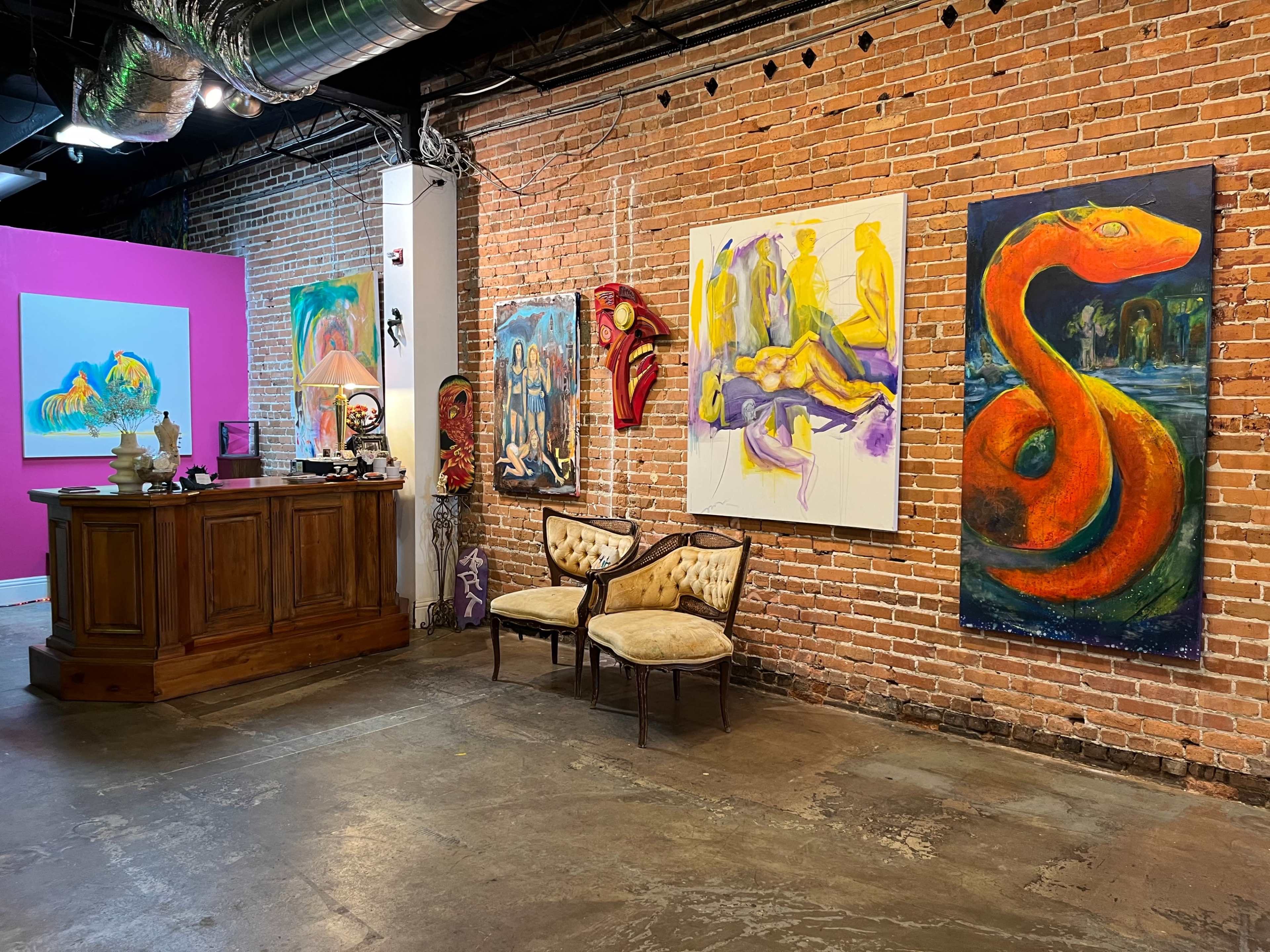 An art gallery with brick walls featuring various colorful paintings, a wooden reception desk, and a seating area with vintage chairs.