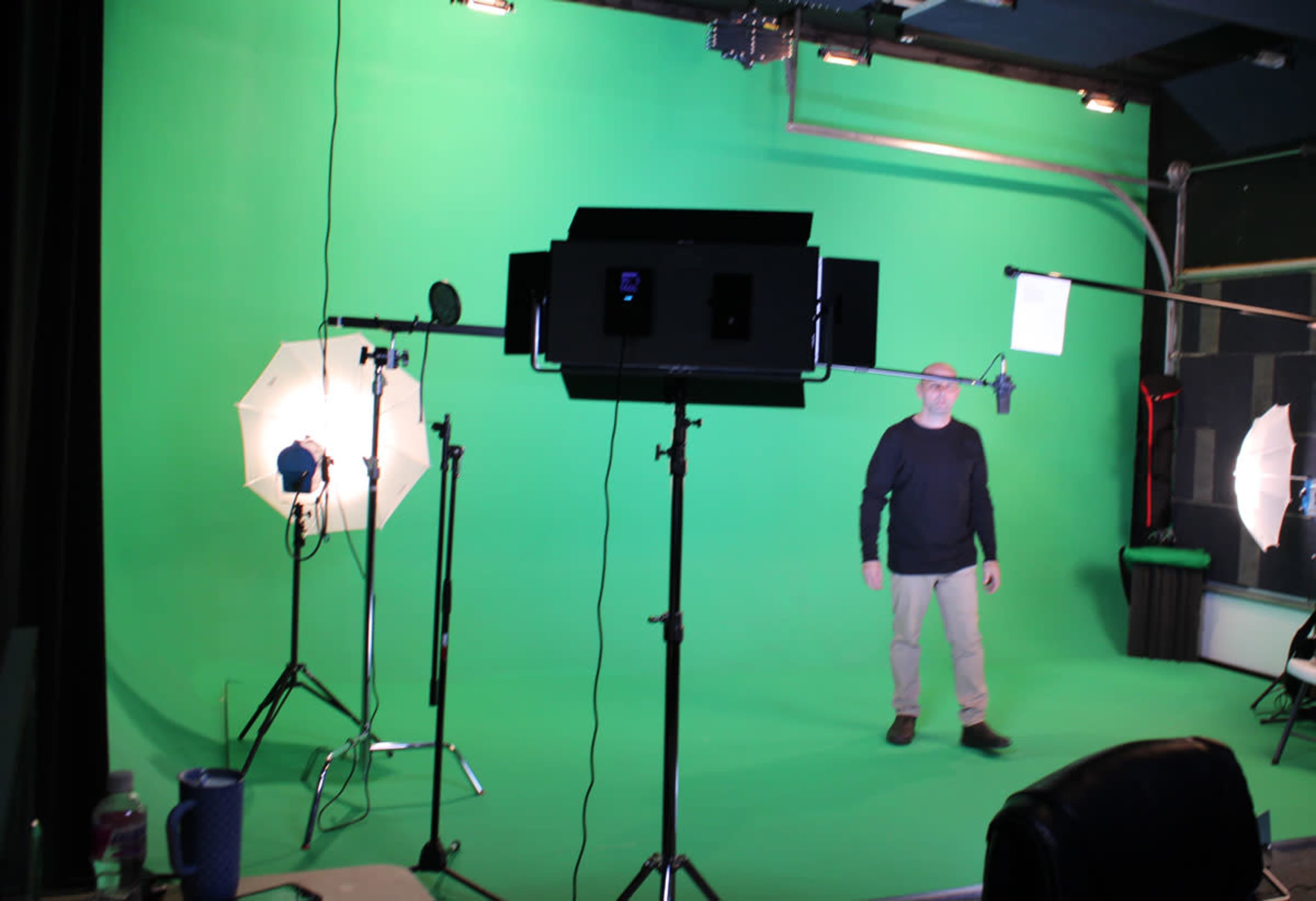 A man stands in a green screen studio surrounded by lighting equipment and a microphone setup.