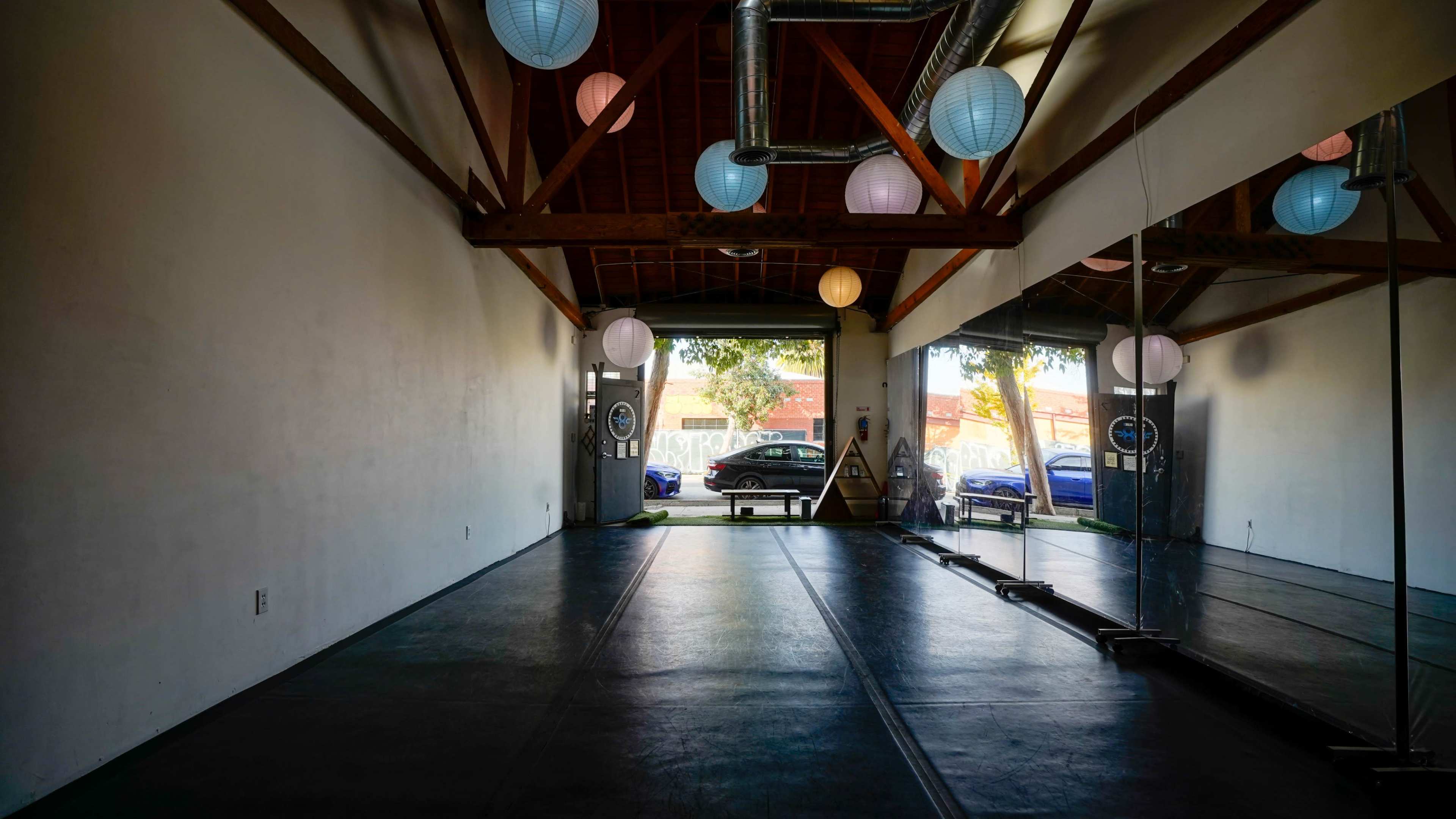 A spacious interior of a studio with large mirrors, colorful lanterns hanging from the ceiling, and windows revealing a view of parked cars outside.